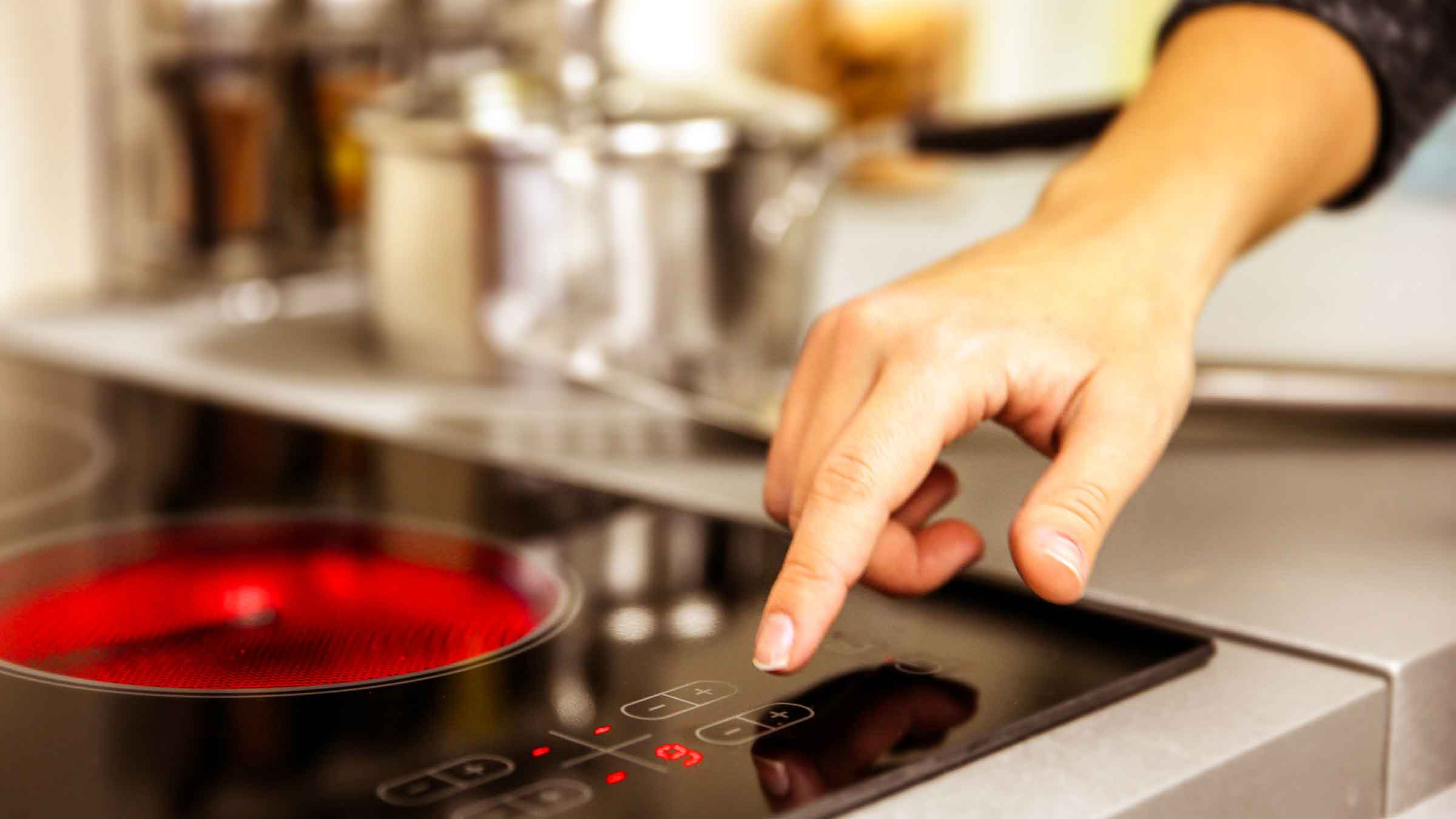 Corner of electric stovetop, bottom right ring is turned on and glowing red, a hand is reaching out to adjust the temperature gauge which is set to nine.
