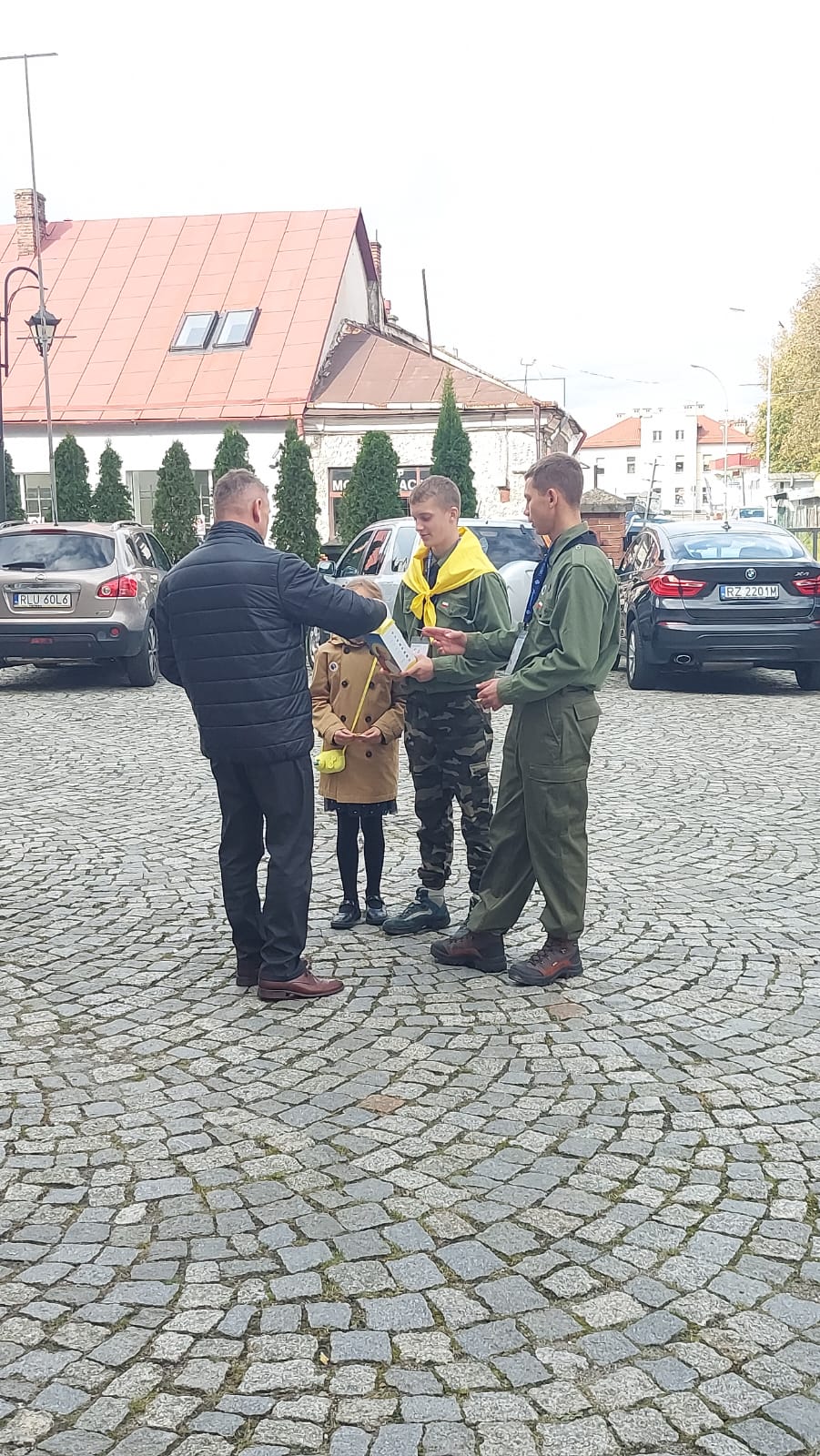 Dzień Papieski | Dwóch chłopców w harcerskich mundurach kwestuje z puszką stojąc na brukowanym placu. Mężczyzna z dzieckiem wrzuca datek do puszki. W tle samochody i zabudowania..jpg