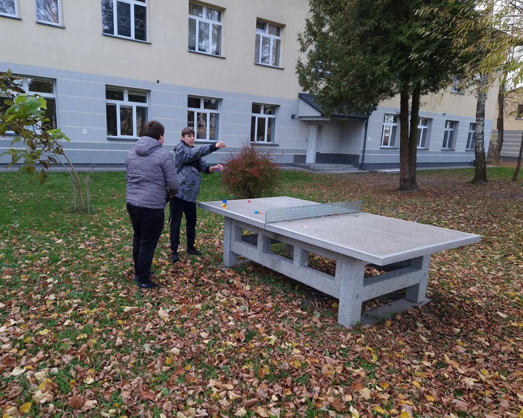 Cykl zajęć z programu „Fajna ferajna”  | Dwie osoby stoją przy kamiennym stole do ping-ponga. Jedna z nich rzuca na stół kolorowe, plastikowe elementy. W tle ściana budynku.jpg