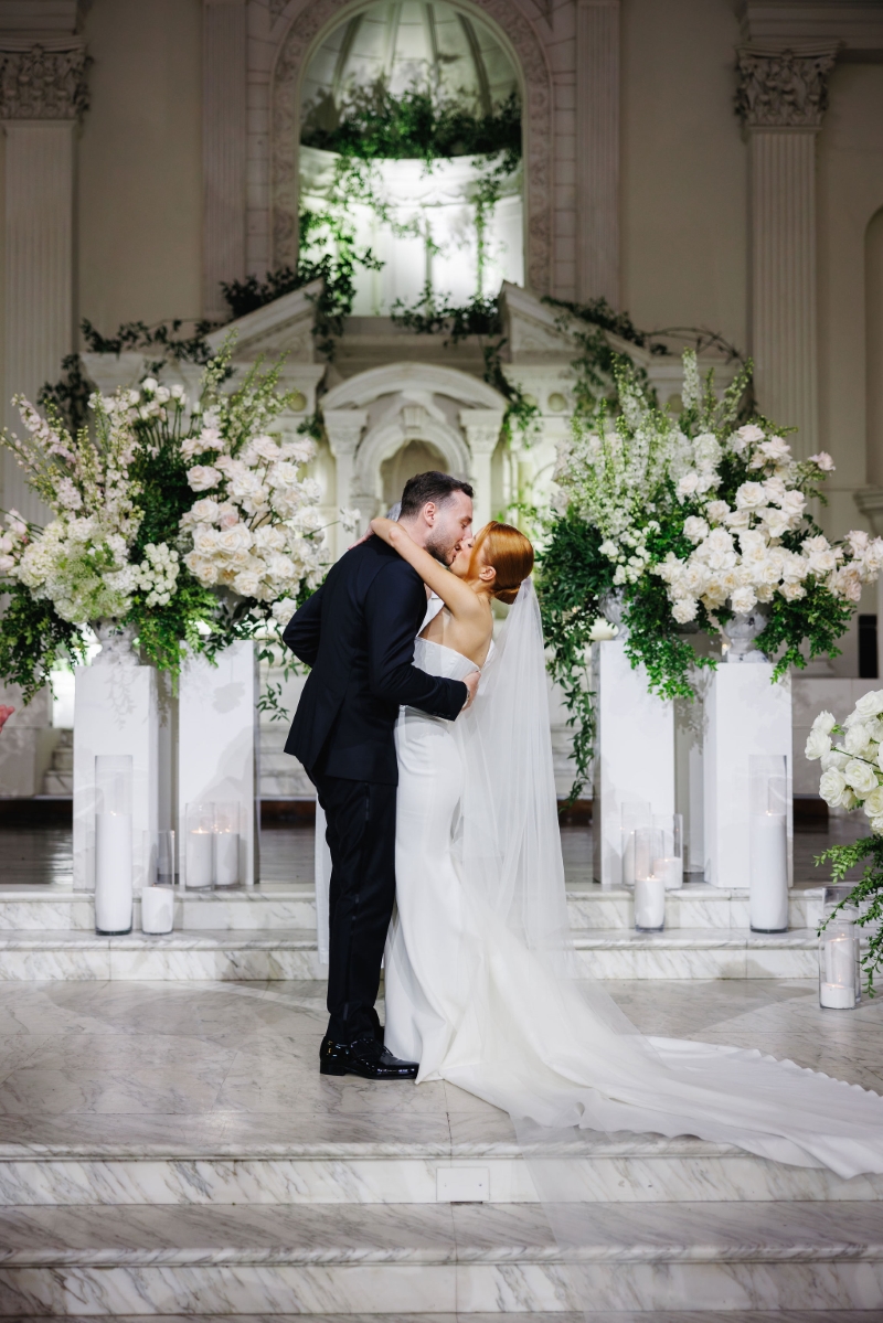 a bride and groom kiss at their wedding ceremony. they are surrounded by white roses