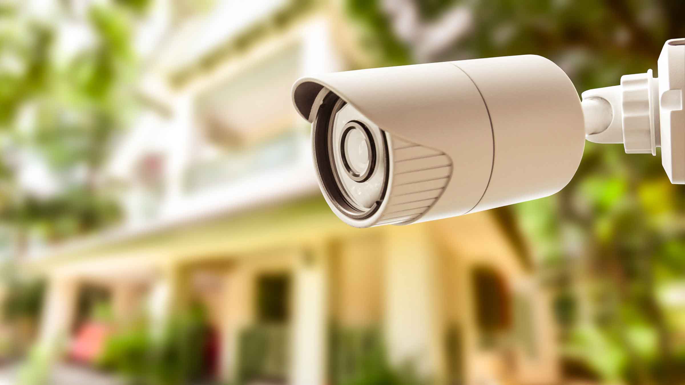 Surveillance camera pointed outward, with blurred home in the background surrounded by greenery.