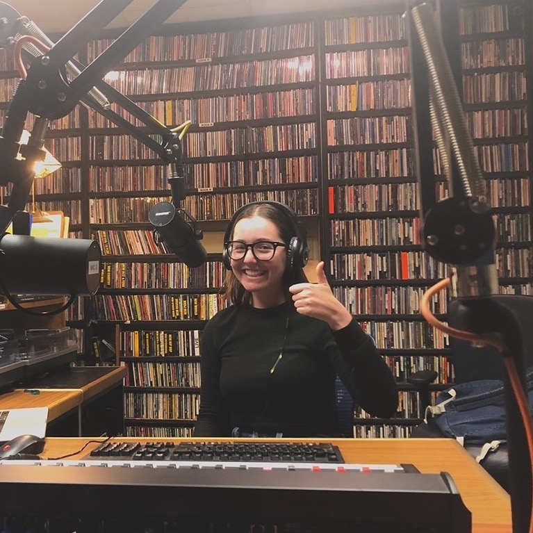 Radio host smiling and giving a thumbs up in a studio with shelves of CDs in the background