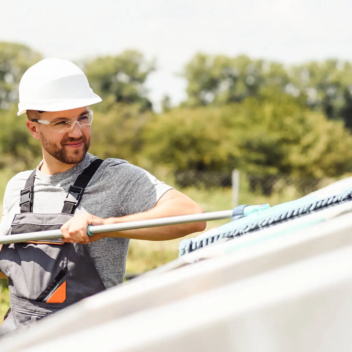 Person with safety helmet and goggles uses a long broom to wash solar panels. In the background is a green forest.