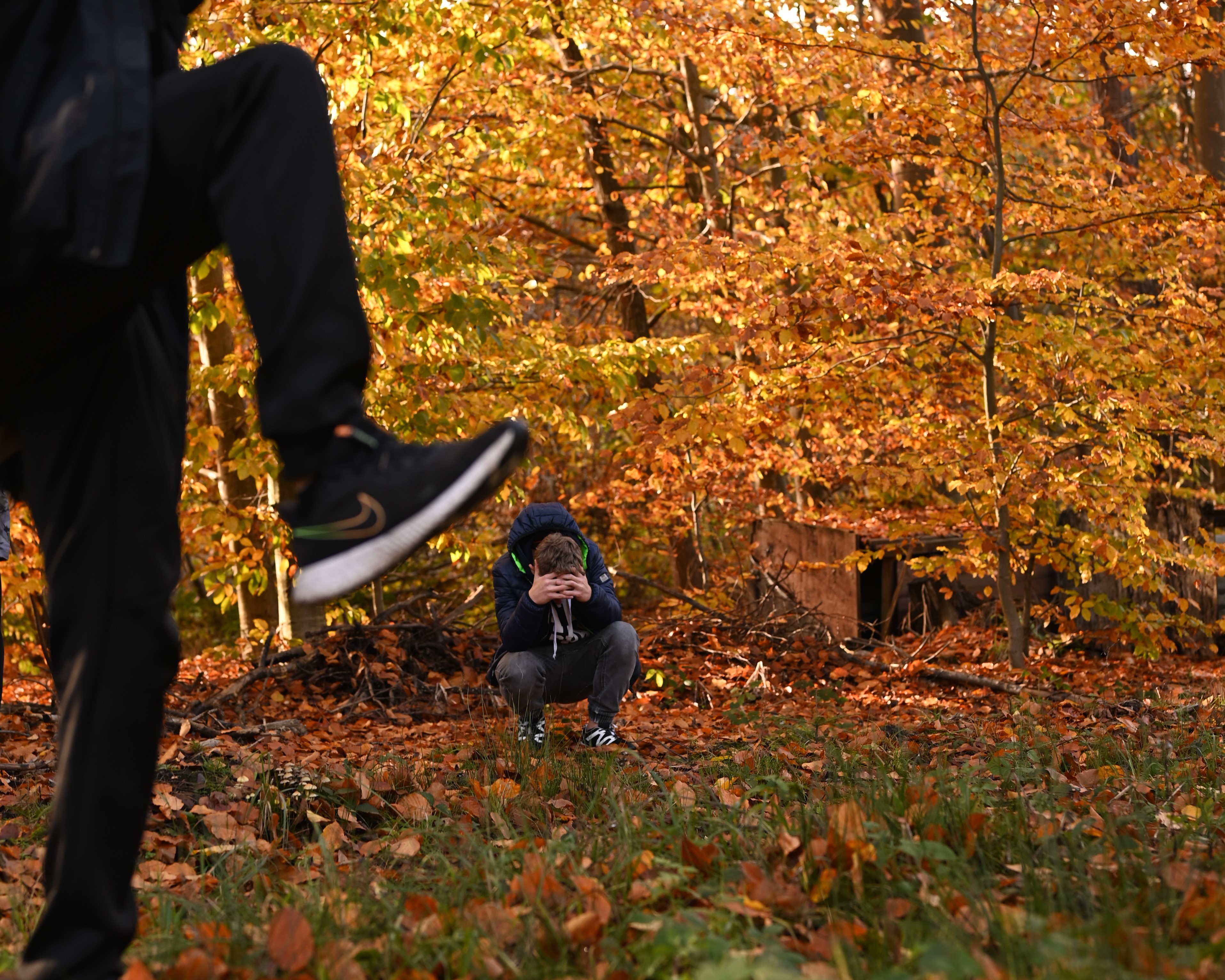 Cykl zajęć z programu „Fajna ferajna”  | Zdjęcie z użyciem perspektywy. Mała sylwetka kucającego, skulonego chłopca na tle jesiennych drzew. Na pierwszym planie ogromne nogi z uniesioną stopą w bucie, jakby ktoś chciał rozdeptać kucającego.JPG