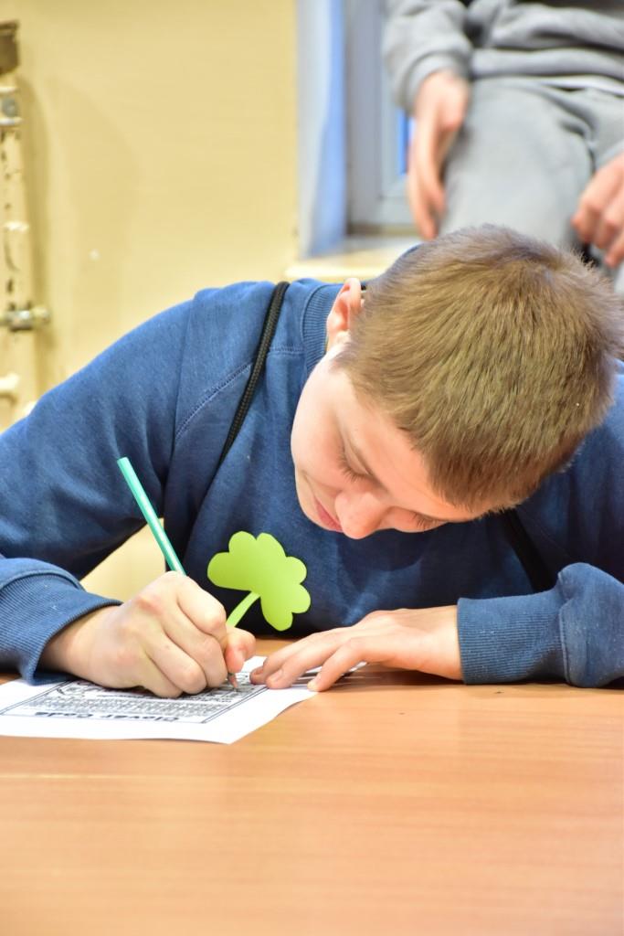 Dzień Św. Patryka  | Chłopiec w szarej bluzie z dużą zieloną koniczynką siedzi za stołem pochylając się nad kartką papieru. Chłopiec pisze coś ołówkiem.JPG