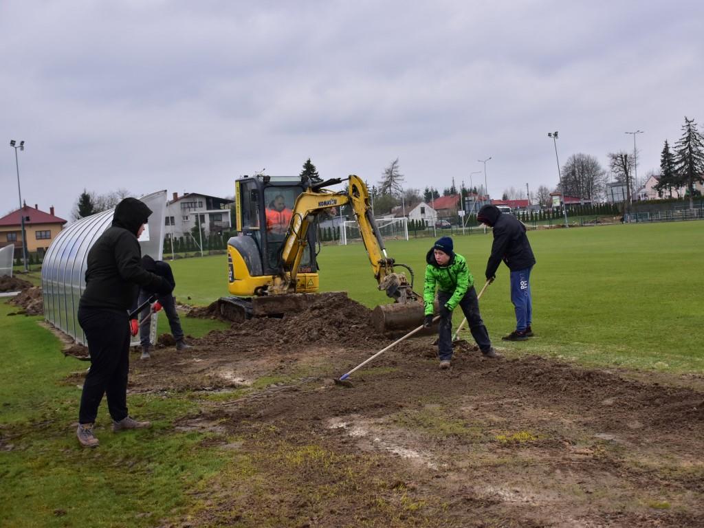 Porządki na stadionie | Cztery osoby ubrane w kurtki grabią rozrytą ziemię na murawie boiska. Za nimi niewielka zółta koparka.JPG