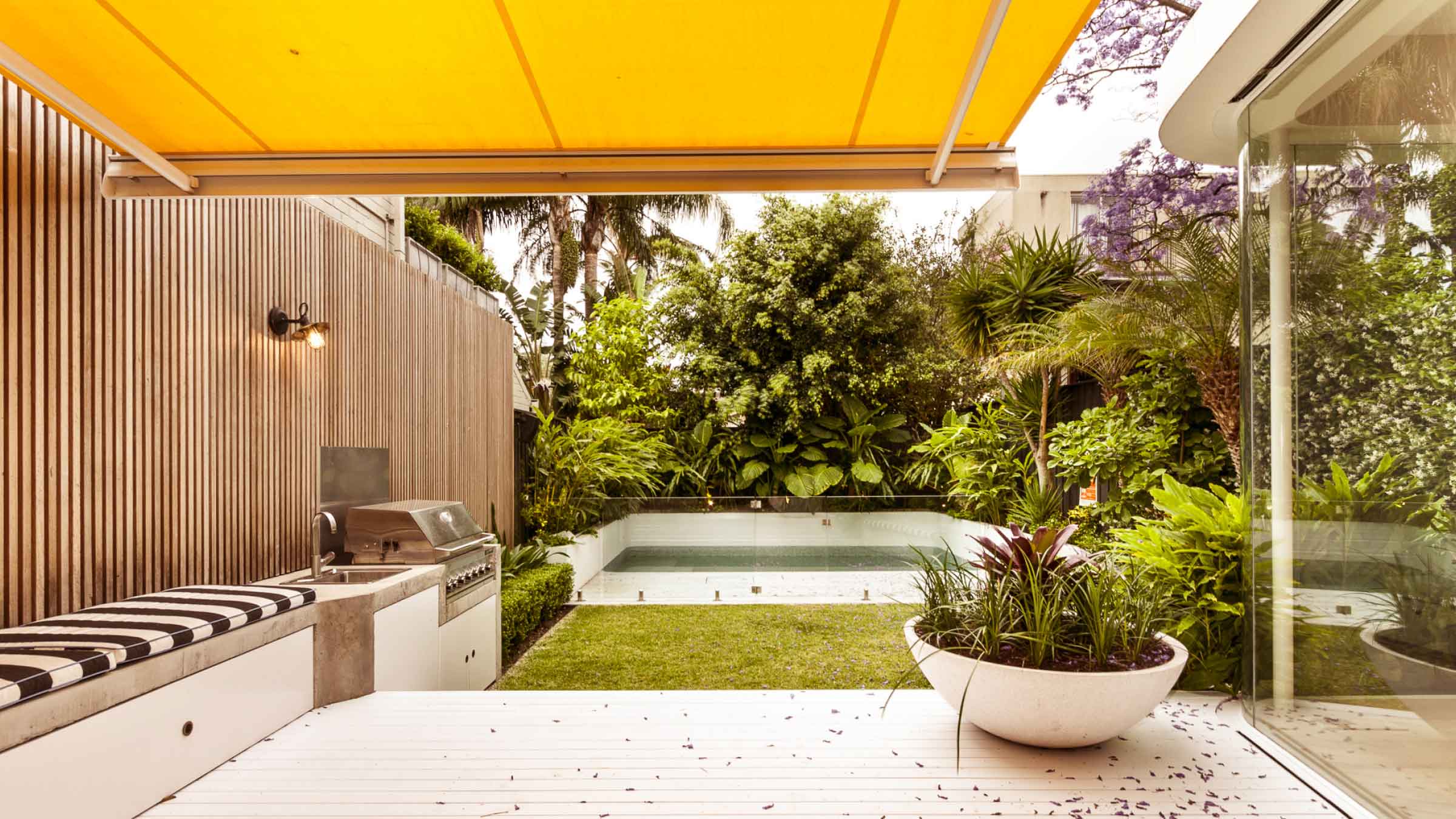 Compact backyard with bbq, seating and wooden paneling on the left. Towards the back of the garden there's a small pool surrounded by luscious green plants. A bright yellow awning covers the entertaining section.