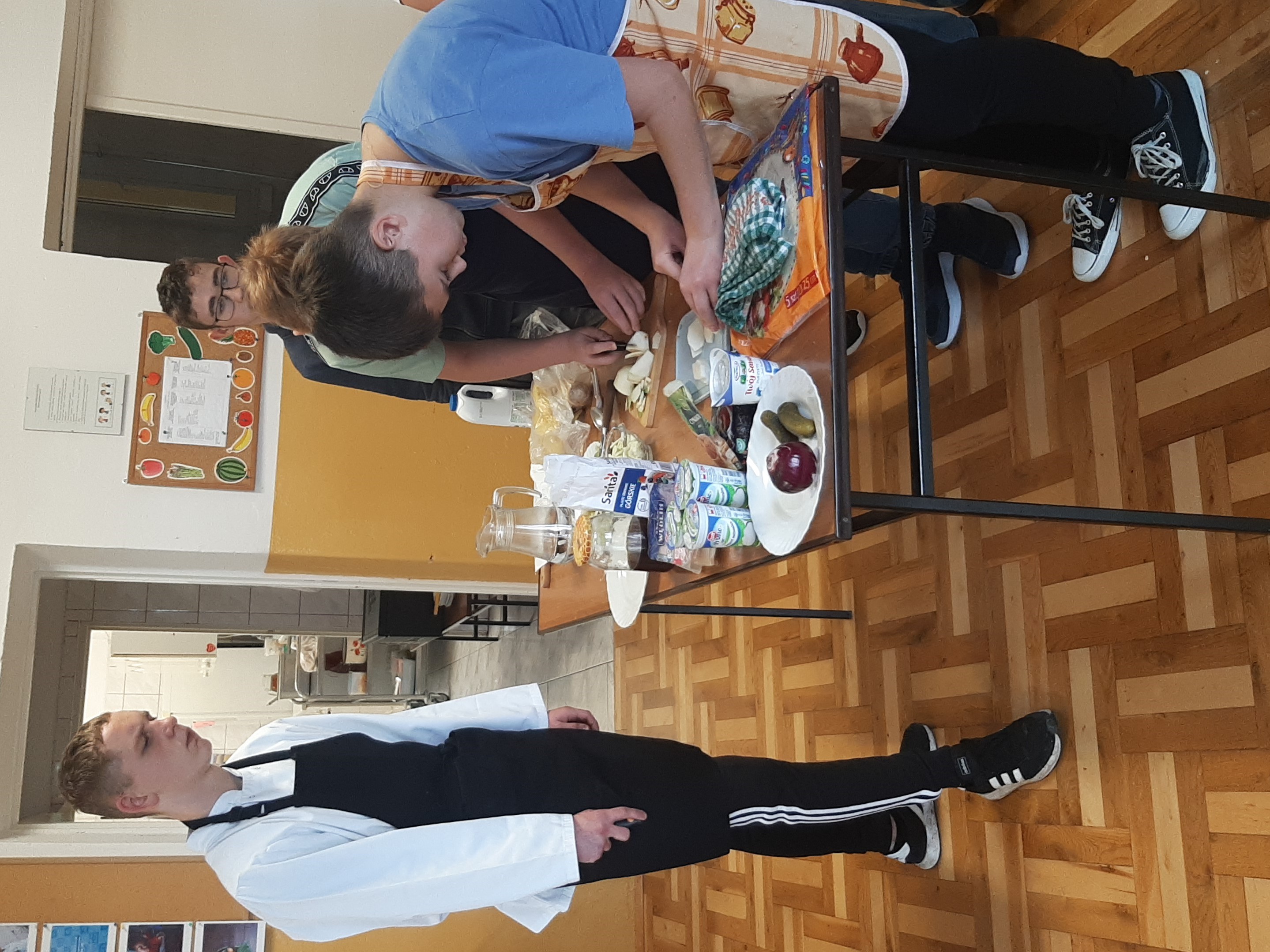 Warsztaty kulinarne | Wychowanek MOW stoi przy stole, na którym leżą produkty spożywcze i nadzoruje przygotowanie przekąsek przez uczniów szkoły podstawowej..jpeg