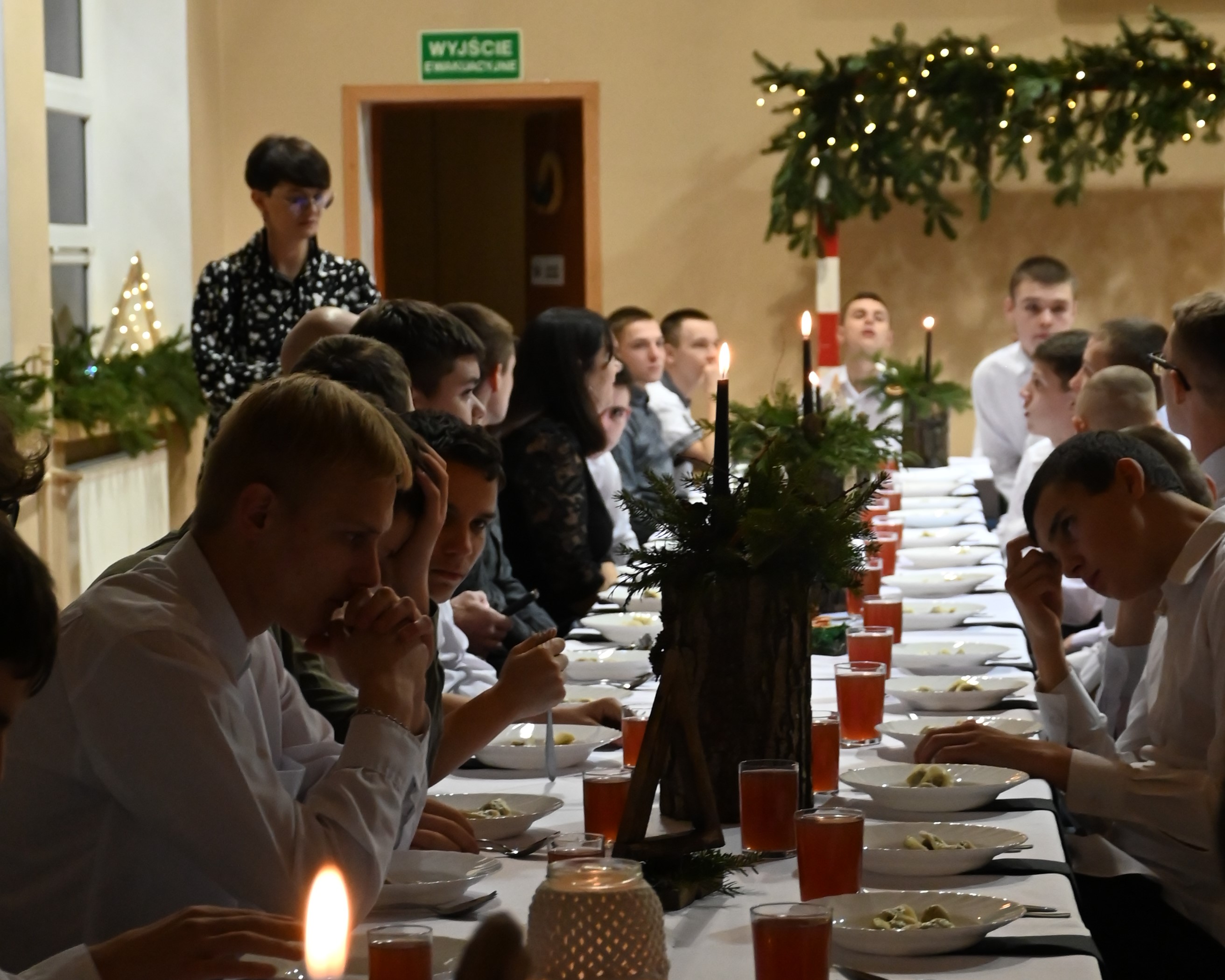 Wigilia w Ośrodku | Zdjęcie zrobione z perspektywy długiego stołu z nakryciem do kolacji wigilijnej. Po obu stronach stołu siedzą wychowankowie.JPG