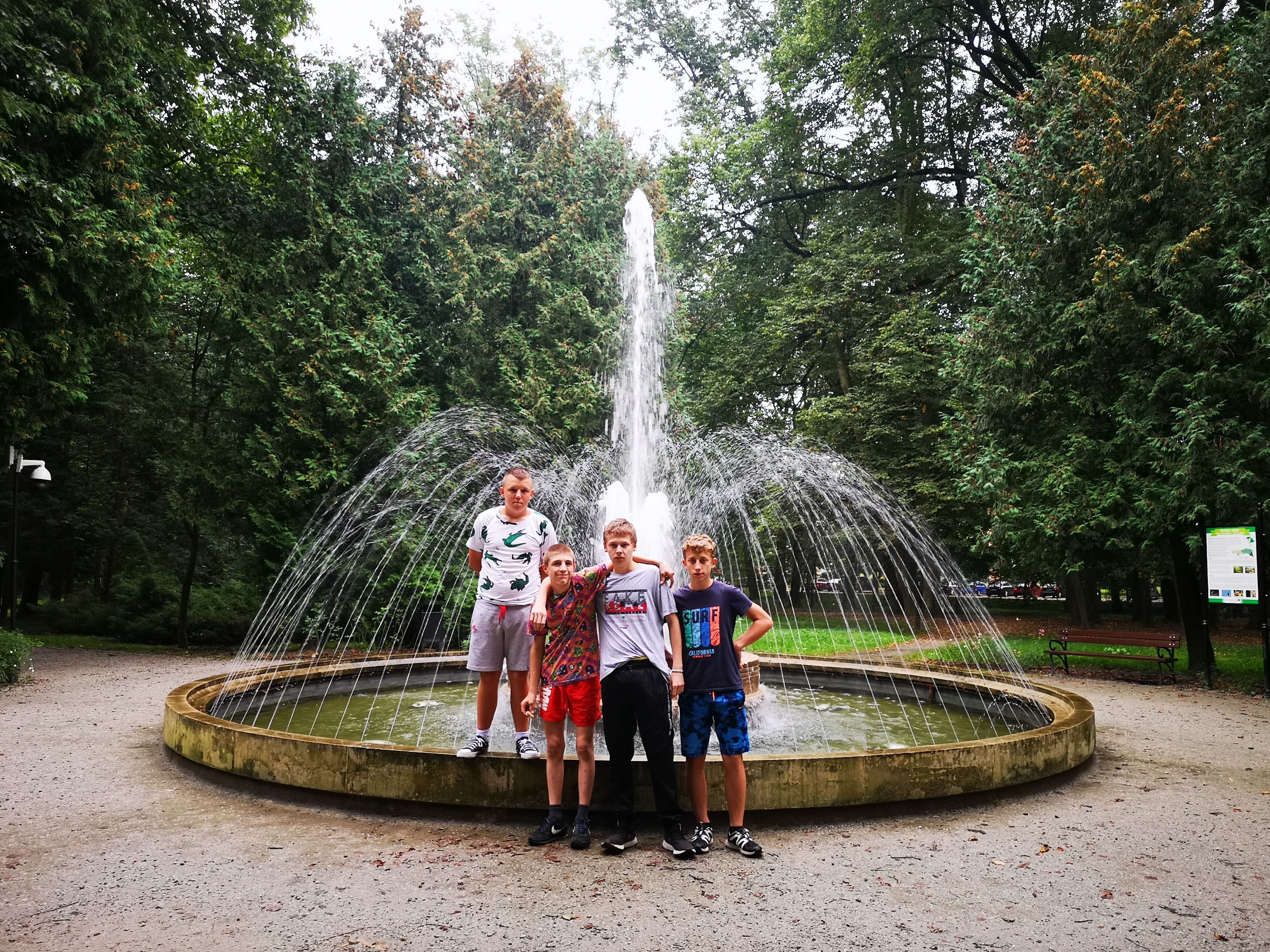 Wycieczka do Jarosławia. | Czterech wychowanków MOW stoi przed okrągłą, tryskającą fontanną usytuowaną na placu w parku, z tyłu gęste, duże drzewa..jpg