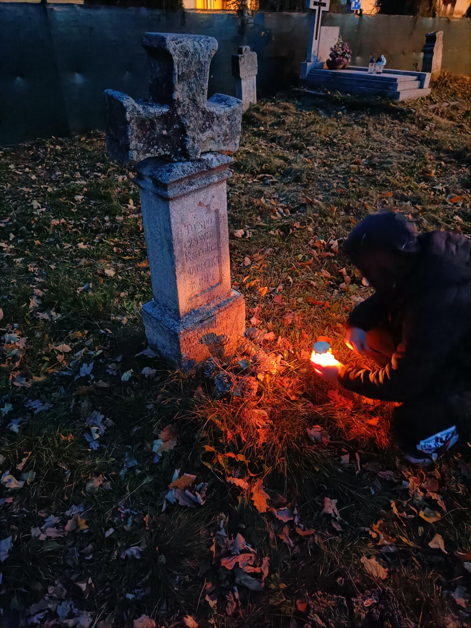 Pamiętamy... | Wychowanek MOW kuca przed kamiennym krzyżem na niewielkim cokole, stawiając na ziemi zapalony znicz. W tle inne mogiły, wokół suche liście. Zdjęcie zrobione o świcie, dość ciemne..jpg