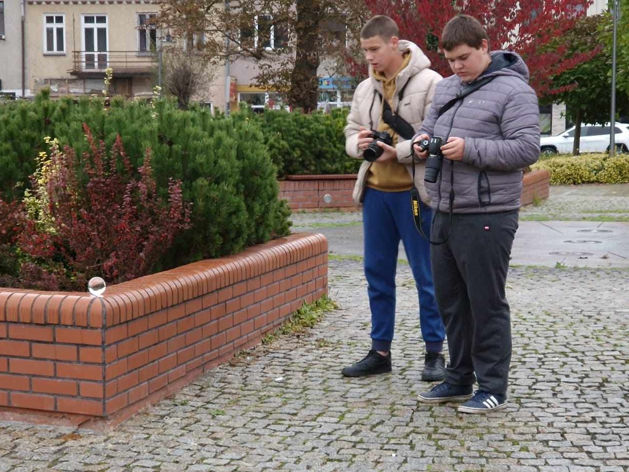 Fotografia jeszcze raz | Dwóch chłopców z aparatami na brukowanym Rynku. Przed nimi ceglany murek, na którym leży szklana kula. Za murkiem krzewy w jesiennych barwach, w tle jesienne drzewa i fragmenty zabudowań..jpg