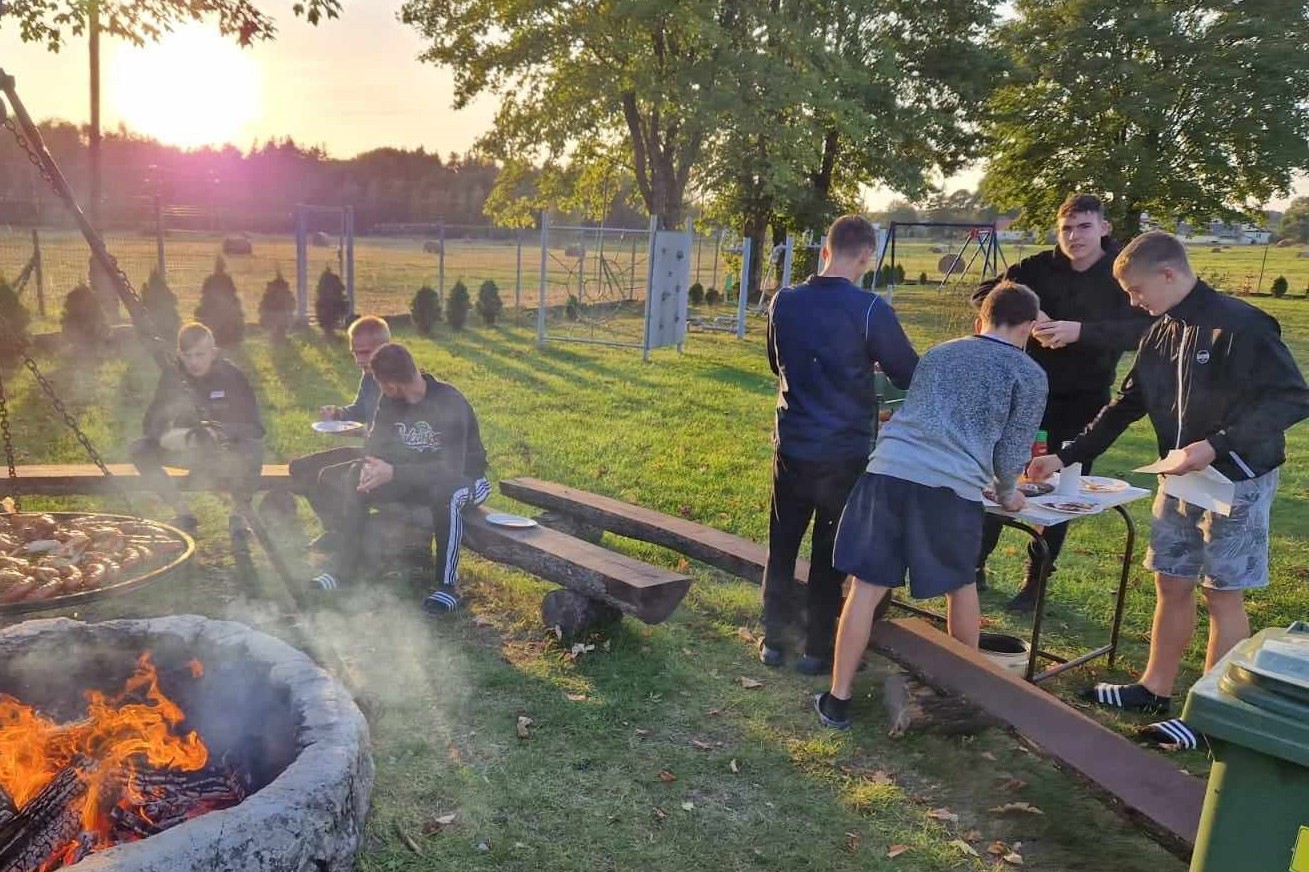 Obóz w Hucie Różanieckiej | Chłopcy przy stoliku biorą z niego talerzyki, sztućce, chleb, przyprawy. Inni siedzą na ławeczkach przy ogniu. W tle zachodzące między koronami drzew słońce..jpg