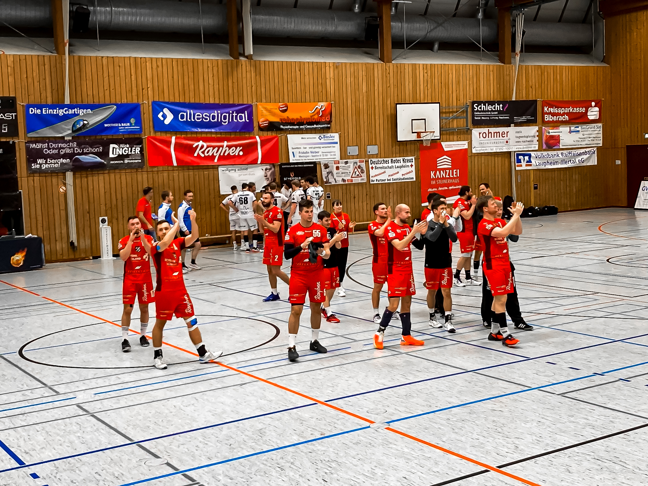 Spieler des HRW Laupheim in roten Trikots jubeln und applaudieren in einer Sporthalle nach einem Handballspiel. Im Hintergrund stehen weitere Spieler und sind Werbebanner an der Holzwand angebracht.