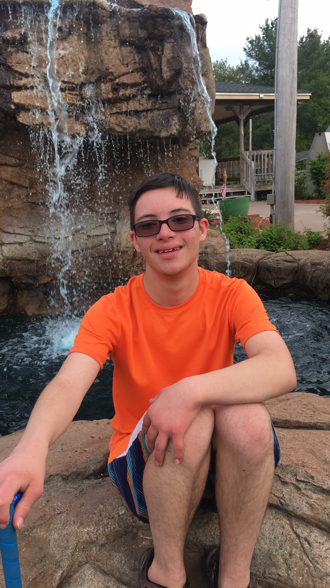 A young man with Down syndrome in a tshirt and sunglasses smiling and sitting in front of a waterfall