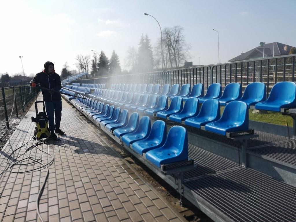 Porządki na stadionie | Chłopiec w kurtce z kapturem myje myjką ciśnieniową plastikowe, niebieskie krzesełka na trybunach.jpg