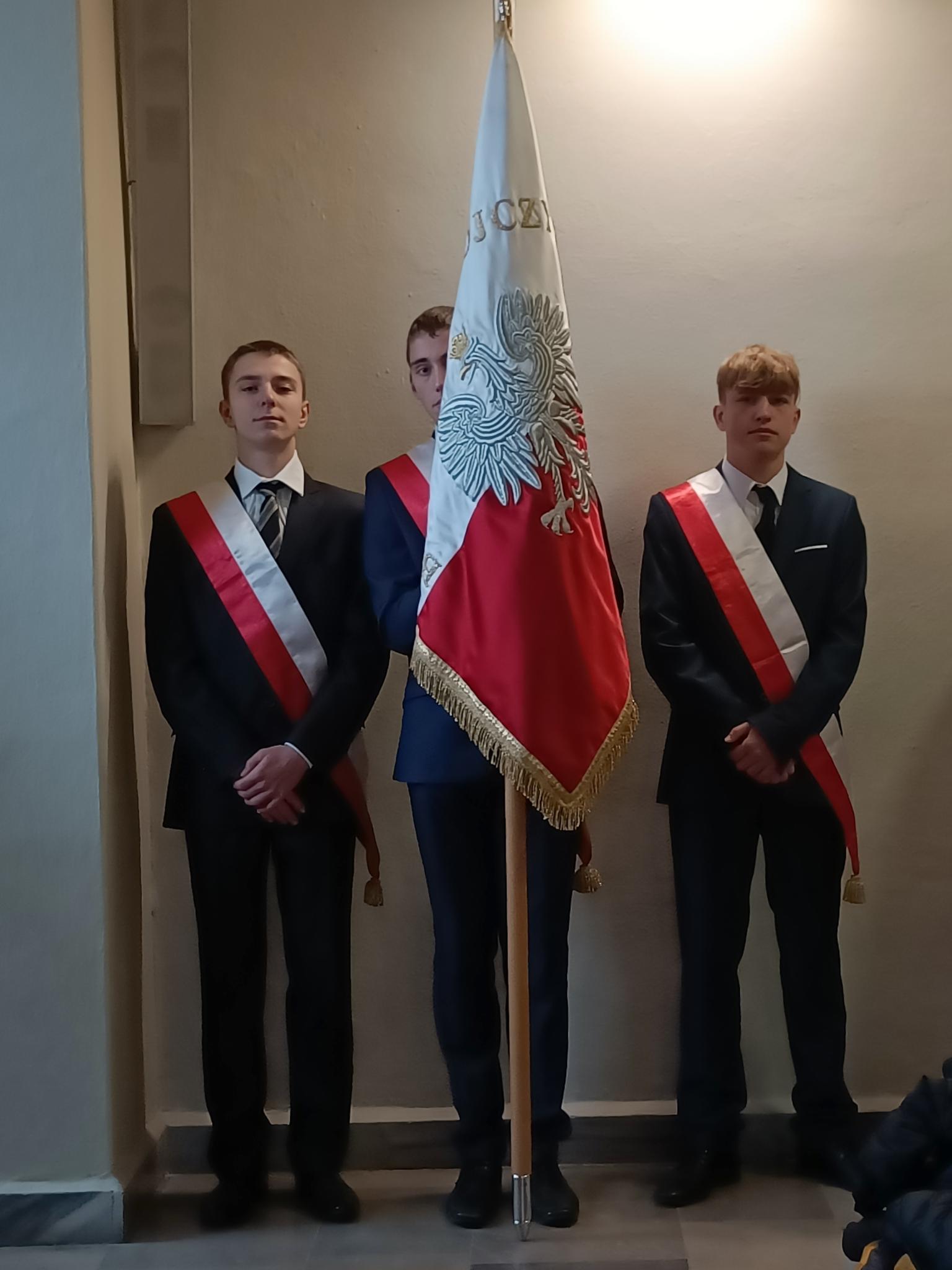 Narodowe Święto Niepodległości | Trzech chłopców w garniturach i biało-czerwonych szarfach stoi trzymając sztandar pod ścianą podczas mszy w kościele..jpg