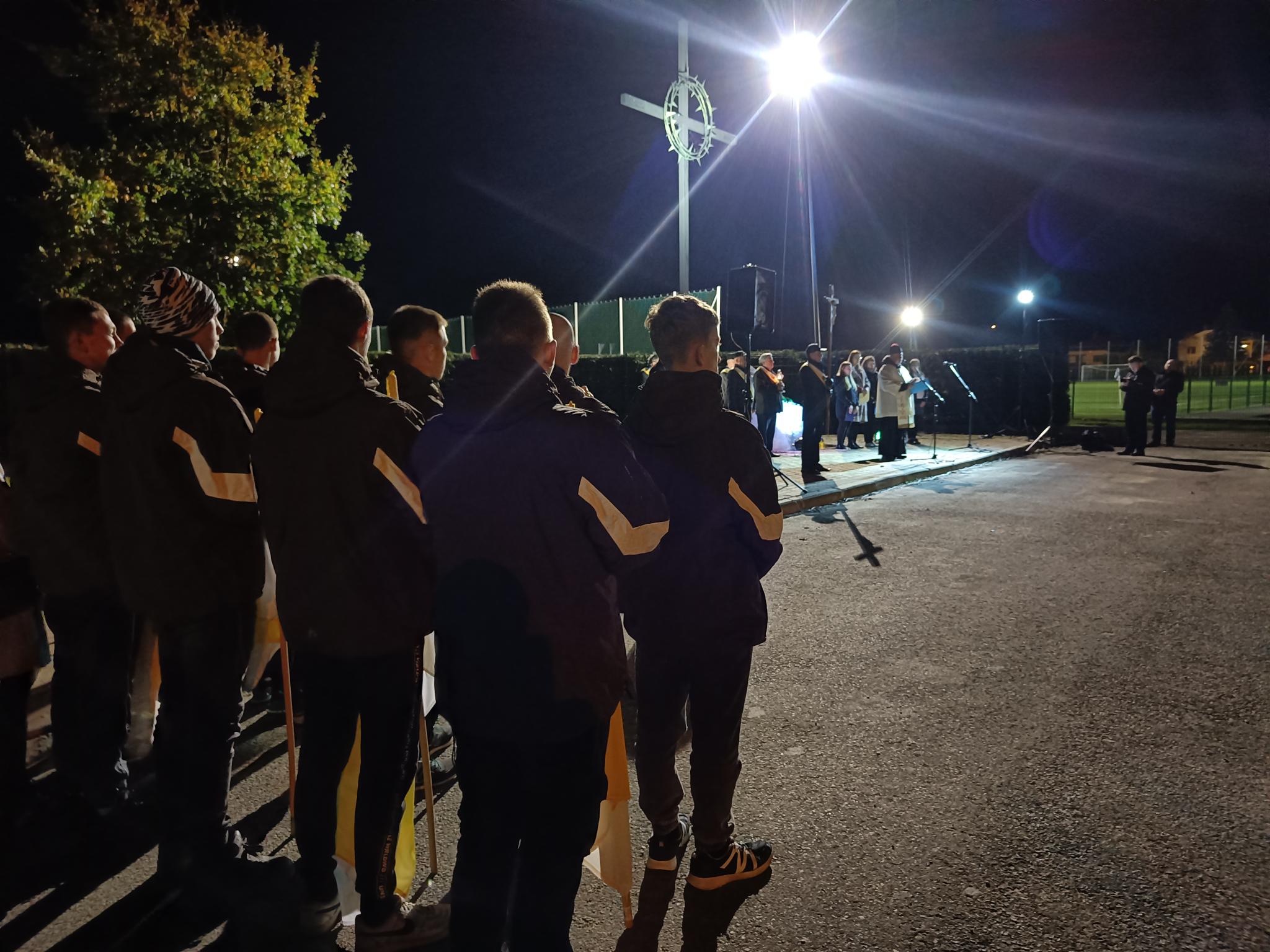 Dzień Papieski | Grupa chłopców w kurtkach stoi tyłem w dwuszeregu, w tle widoczny wysoki krzyż, pod którym odprawiane jest nabożeństwo. Zdjęcie nocne..jpg