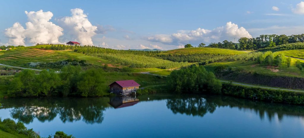 A small house is nestled into the hillside in front of a lake. The background is rolling green hills and a blue sky.