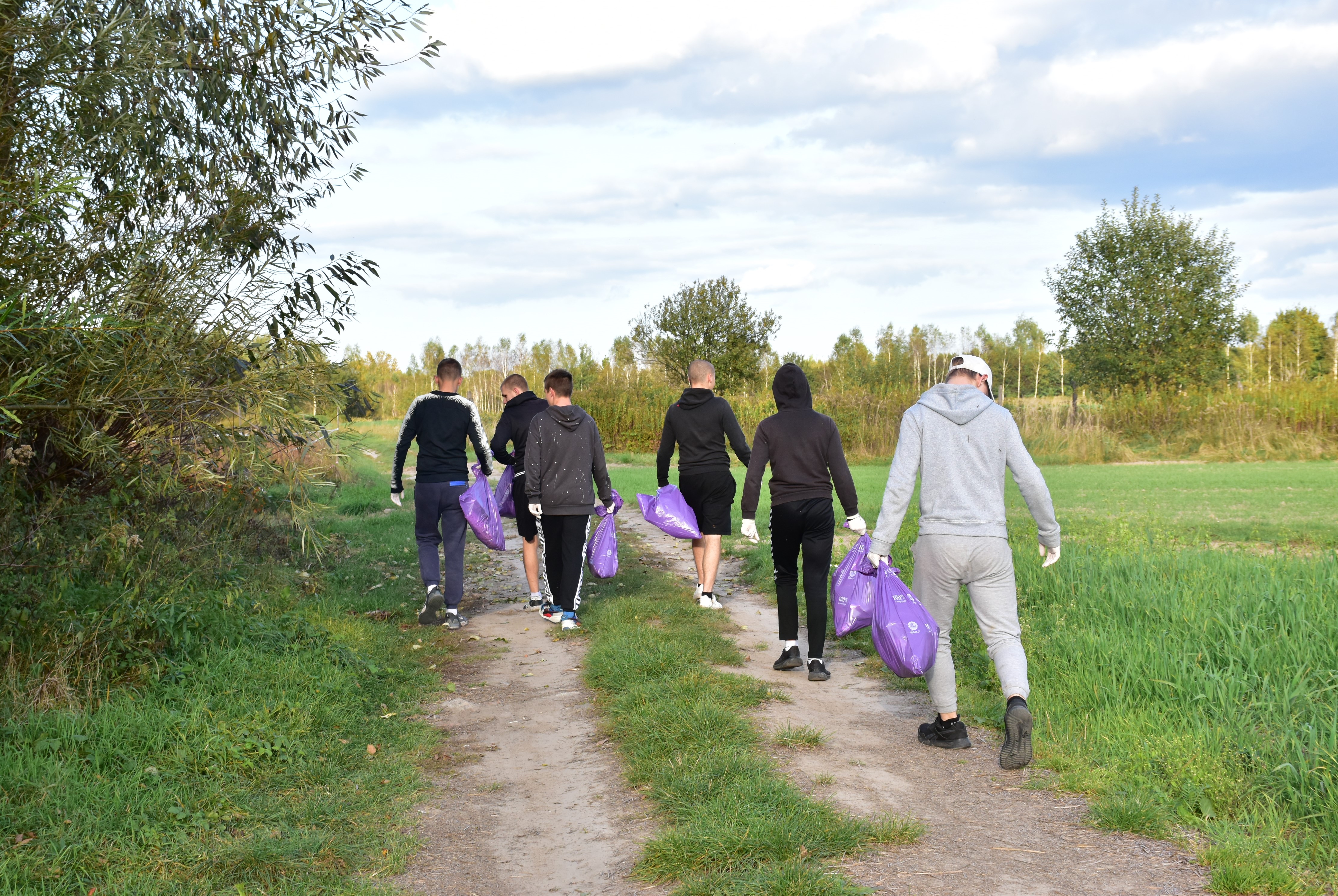 Sprzątamy dla Polski | Grupa chłopców widzianych tyłem idzie po polnej drodze, każdy niesie fioletowy worek. dokoła nich zieleń - trawa, krzewy, pojedyncze drzewa..JPG