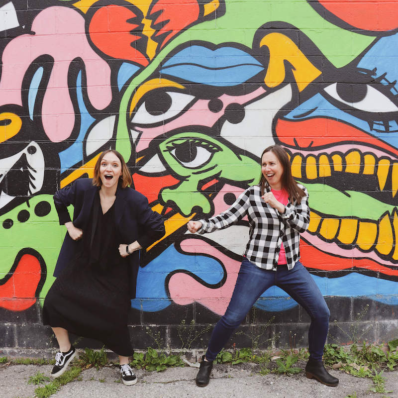 Two female coworkers joking around in front of a colorful mural