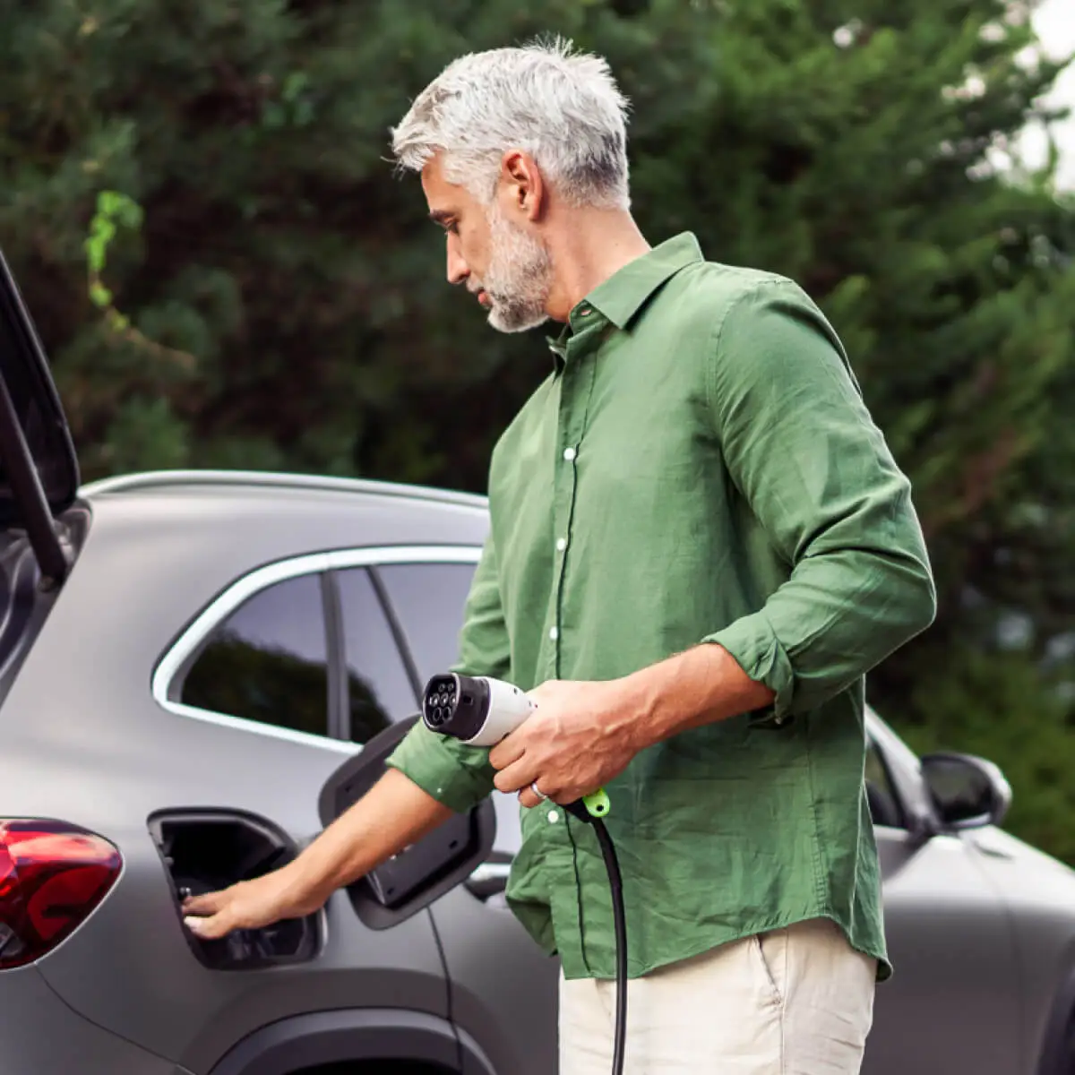 Older man in green linen shirt, opens plug cap on his electric vehicle to begin charging it.
