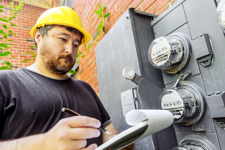 Workman with yellow hard hat and clip board, inspects an electricity meter outside a red brick home.