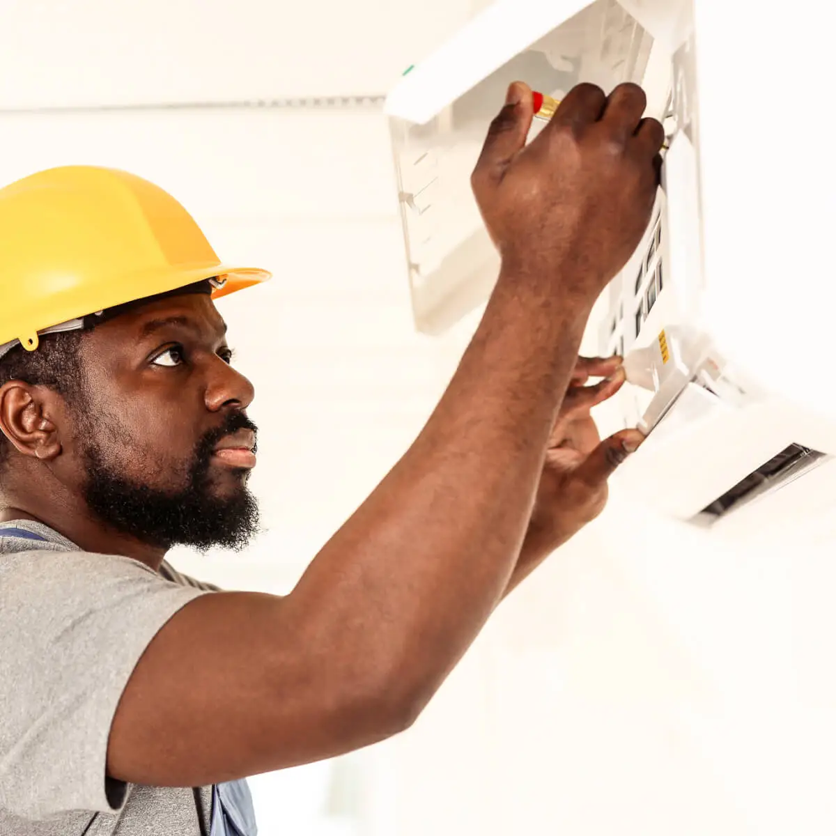 Middle aged man in yellow hard hat and overalls, inspects an air conditioner mounted on a wall.