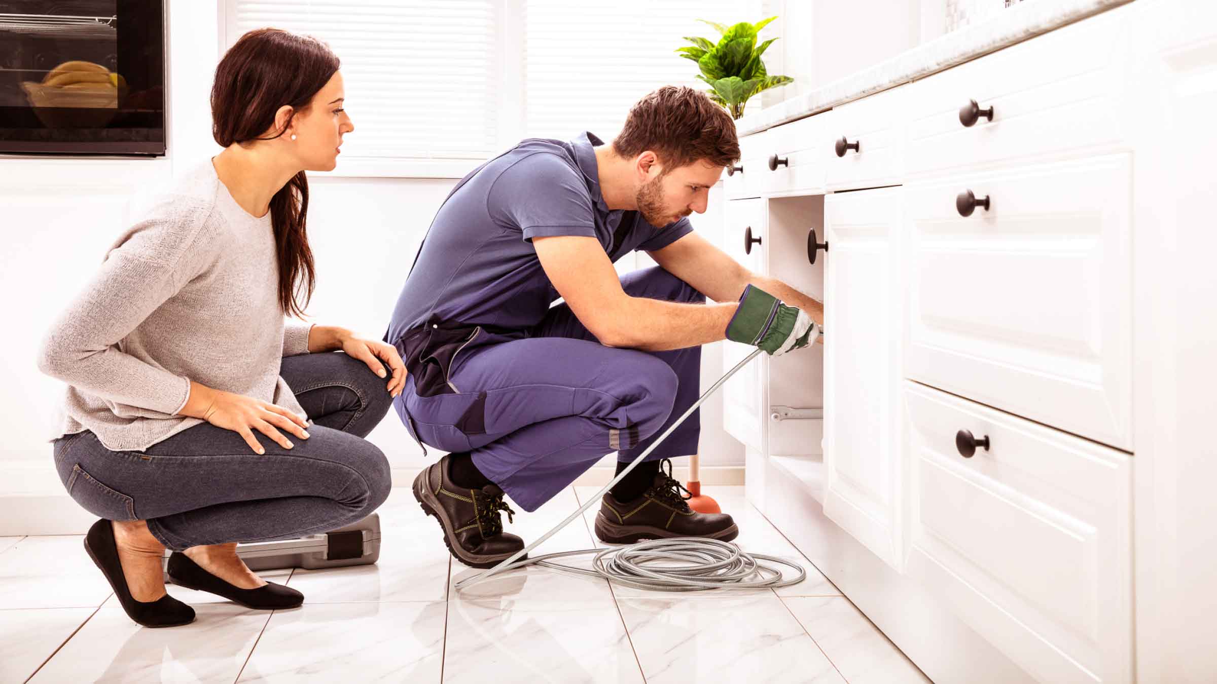 Woman and tradie kneel in front of kitchen cupboard inspecting plumbing.