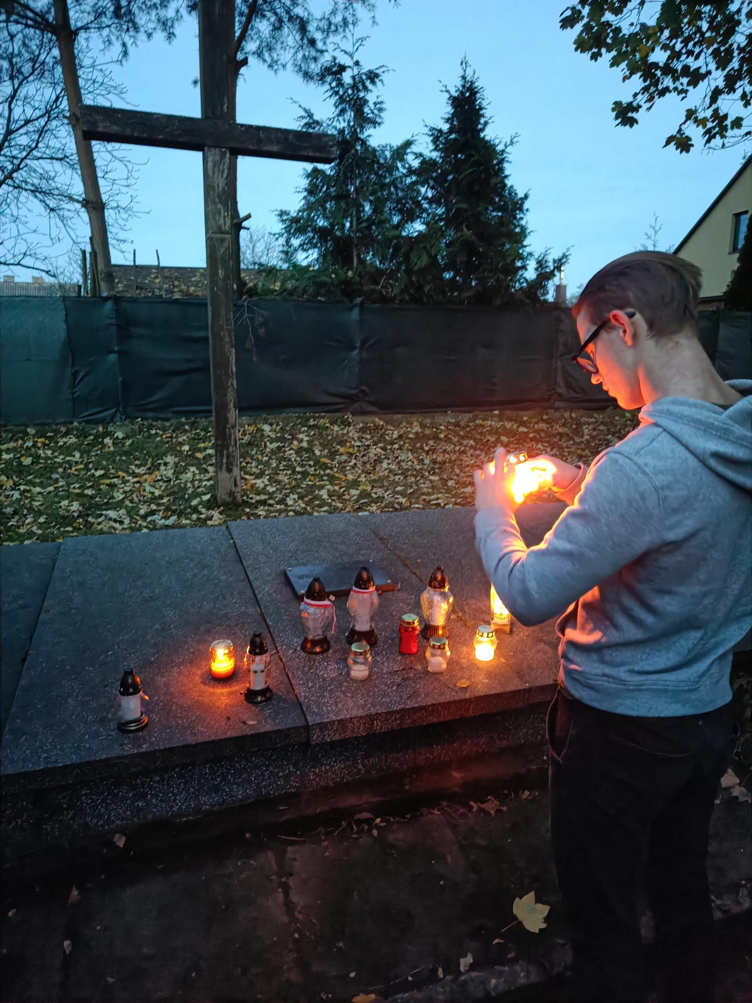 Pamiętamy... | Chłopiec w szarej bluzie zapala znicz przed lastrikową mogiłą. Na płycie palą się inne znicze, za płtyą wbity w ziemię duży drewniany krzyż. Zdjęcie zrobione o świcie, jeszcze ciemnawo na dworze..jpg