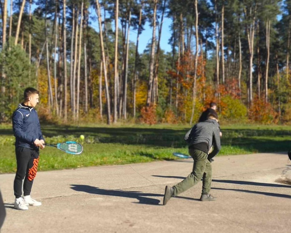 Zajęcia w szkółce leśnej | Dwóch chłopców z rakietkami do tenisa, jeden odwrócony tyłem, na utwardzonym placu, podczas gry. W tle trawa i jesienny las..jpg