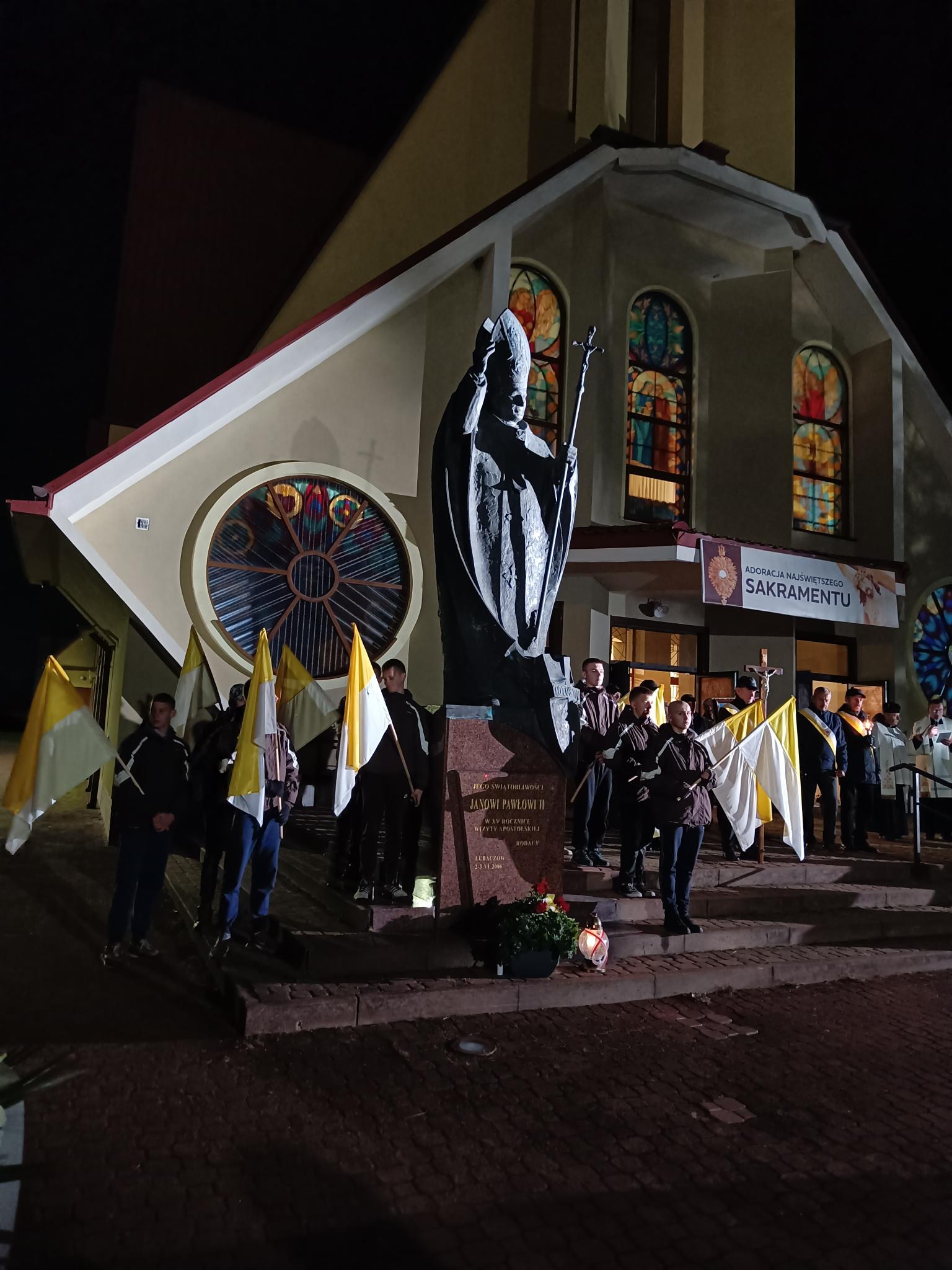 Dzień Papieski | Wychowankowie MOW trzymają wartę honorową przy pomniku Jana Pawła II. Chłopcy stoją po dwóch stronach pomnika, każdy trzyma flagę papieską. W tle fasada kościoła z witrażami. Zdjęcie zrobione nocą..jpg