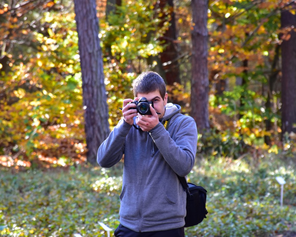 Zajęcia w szkółce leśnej | Chłopiec w szarej bluzie stoi na tle jesiennego lasu przykładając aparat do twarzy i robi zdjęcie fotografującemu..JPG