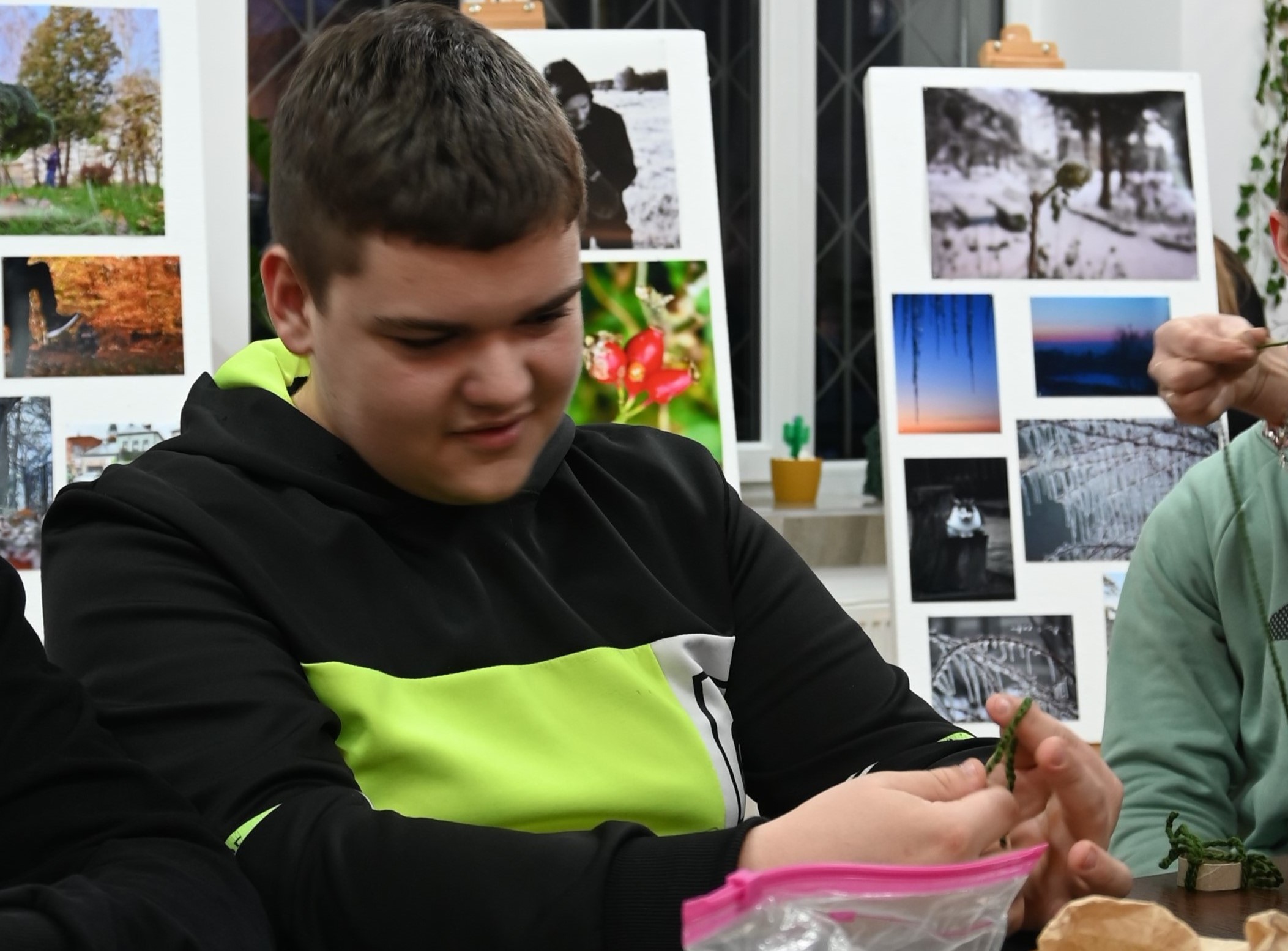 Zakończenie projektu Fajna Ferajna | Chłopiec w czarnej bluzie podczas wiązania na palcach zielonego sznurka. Z przodu fragment foliowej torebki. W tle plansze ze zdjęciami.JPG