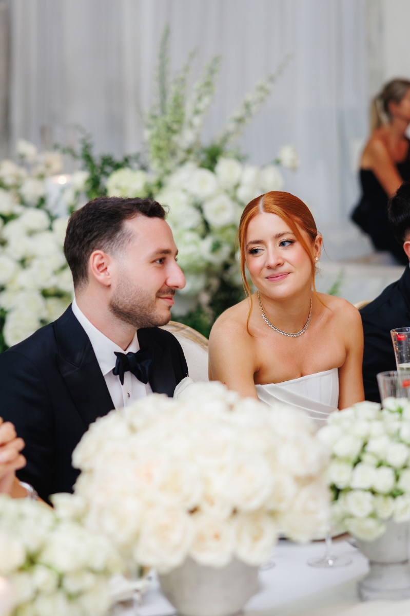 a bride gazes longingly at her groom as they're seated at their table at their wedding reception