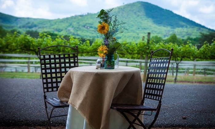 Two empty outdoor chairs are seated at a round table with a white tablecloth. The only thing on the table is a vase with sunflowers in it. The background is lush green vegetation at the foot of a mountain.