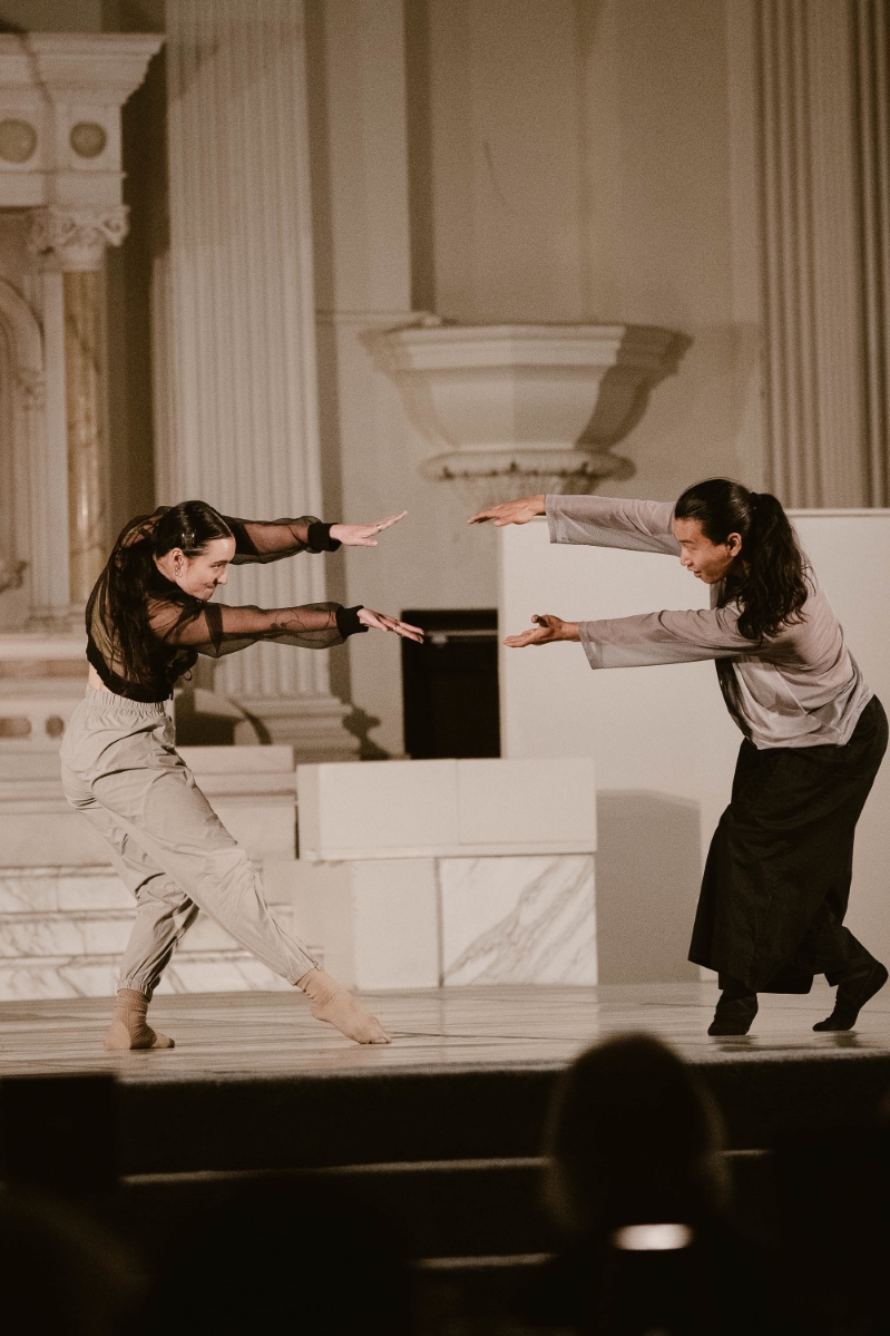 two dancers dressed in neutral colors pose together with their arms outstretched toward each other