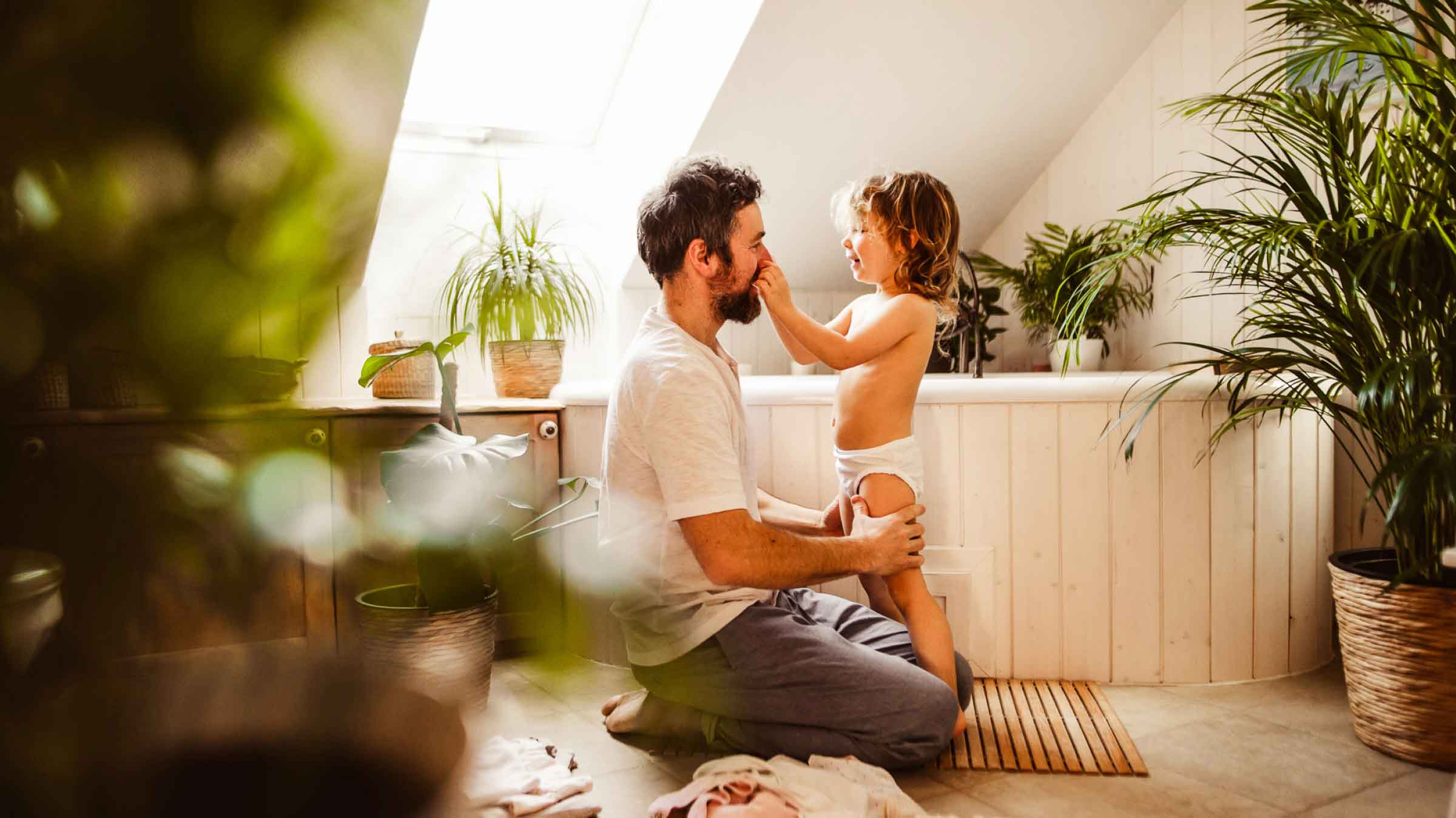 Parent kneels beside bath tub while a child tweaks his nose. Green house plants are dotted around the bathroom.