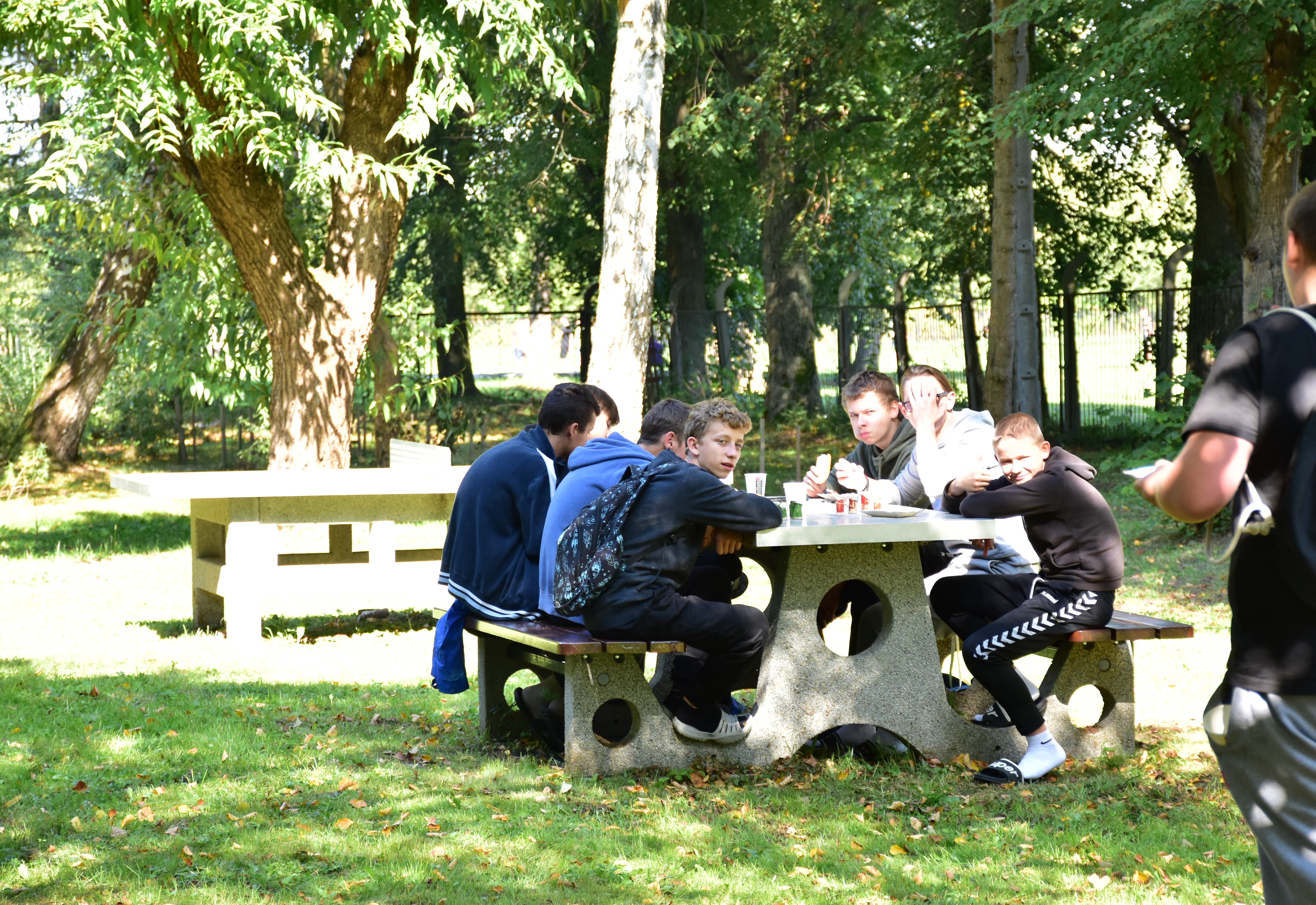 Uroczyste podsumowanie projektu „Dostępna Szkoła” | Wychowankowie MOW siedzą przy kamiennym stole na ławeczkach ustawionych na trawie i jedzą pieczone kiełbaski i szaszłyki. Za nimi kamienny stół do ping-ponga oraz krzewy i duże drzewa..JPG