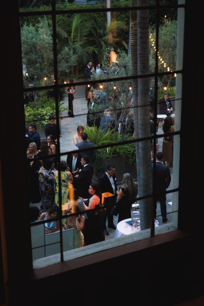 looking through a large cathedral window down into the courtyard where wedding guests mingle and drink at a wedding reception