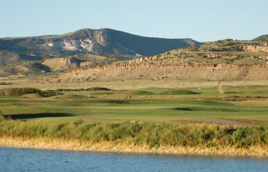 A far-away view of a few holes of a golf course. In the foreground there is a lake and the background is mountains
