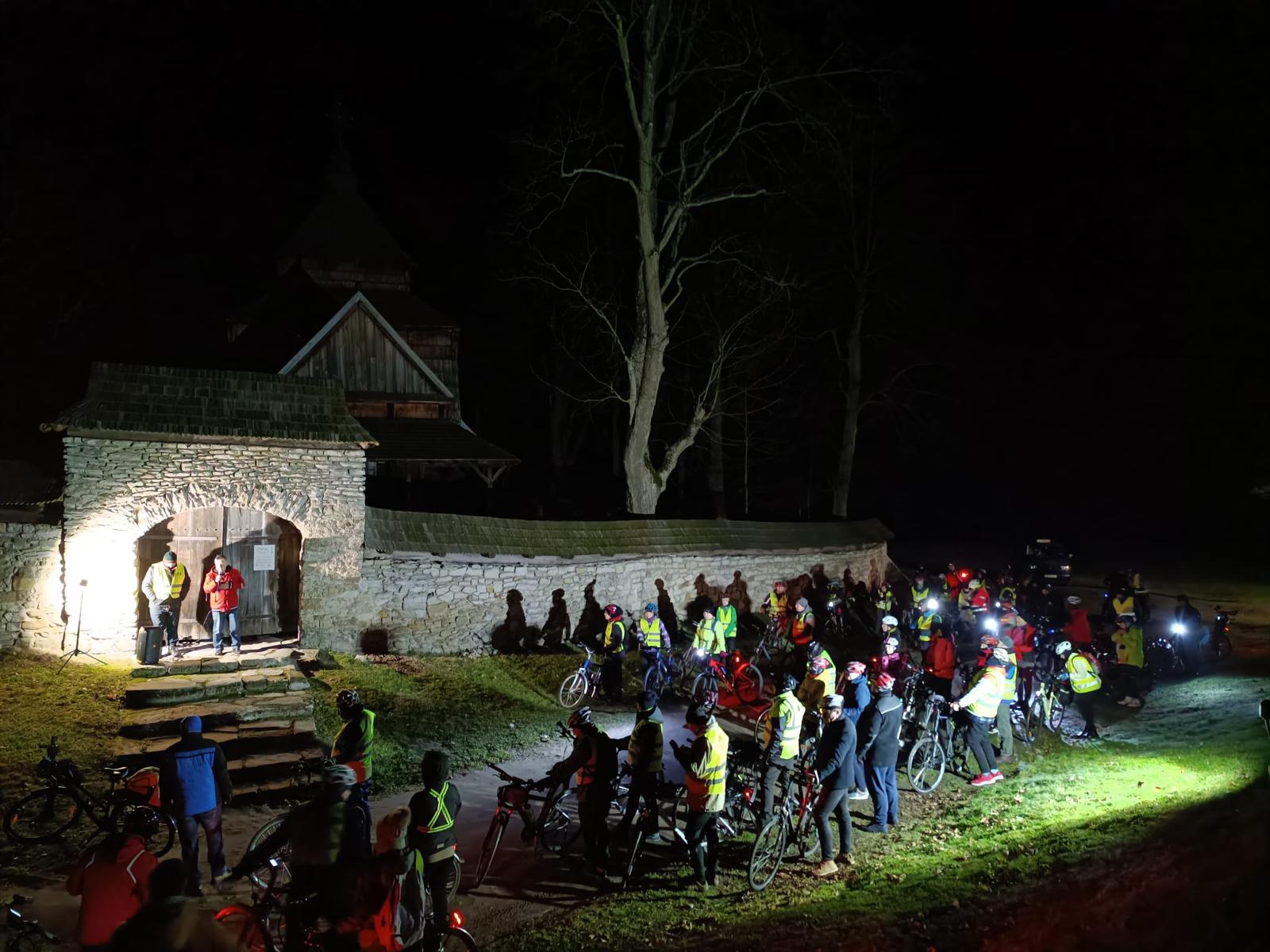 Rowerowa Droga Krzyżowa | Kolarze modlą się stojąc przed bramą wejściową na plac cerkiweny. W tle za murem widoczny fragment cerkwi i ogromne drzewo. Zdjęcie nocne.jpg