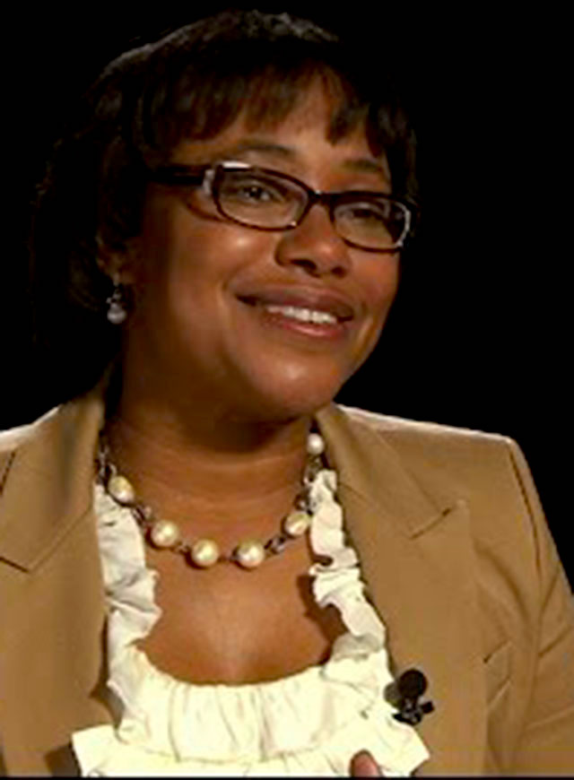 Paula Hammond seated in front of a black background for an on camera interview, smiling.