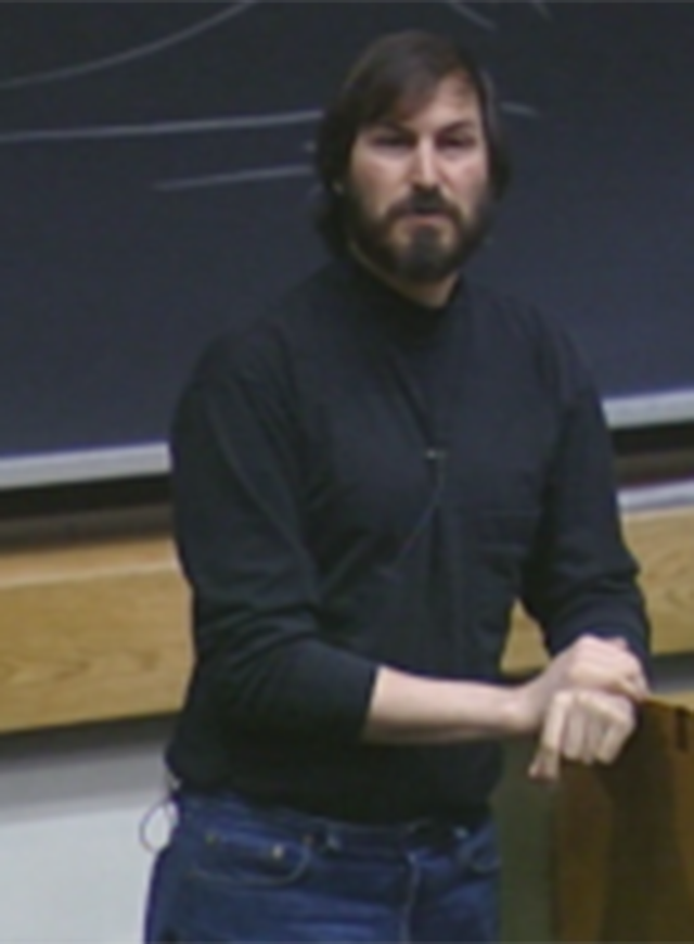 Steve Jobs speaking in classroom, leaning on the podium and standing in front of chalkboard
