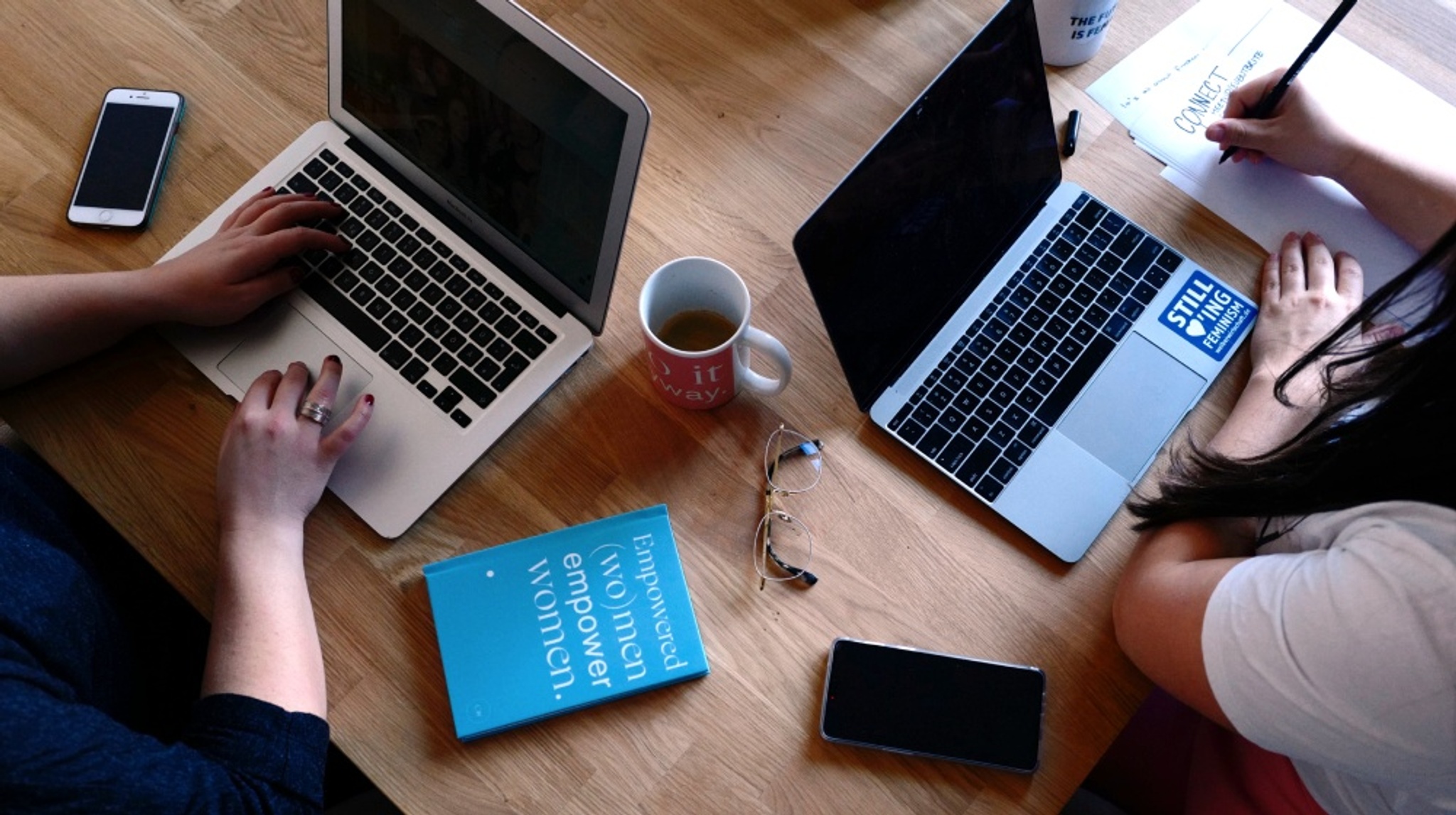 View on a desk from above: two women sit on a desk next to each other with their phones and coffee cups besides their laptops. The scene prominently shows a journal with the text "Empowered (wo)men empower women." A sticker on one laptop says "Still loving Feminism".