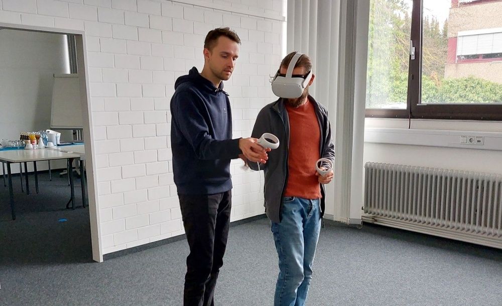 One man uses VR glasses, another man helps him