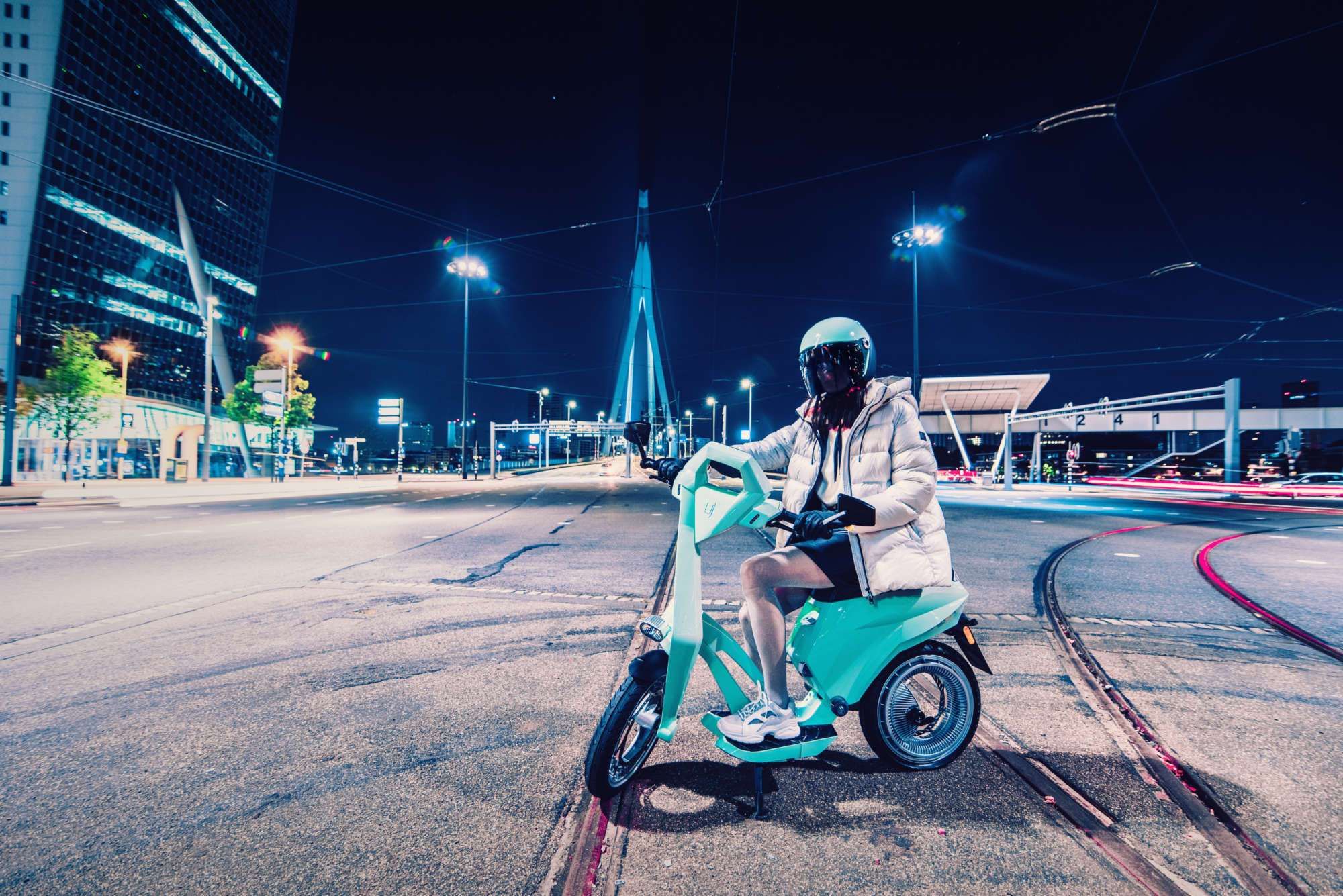 Woman with winter jacket and motorcycle helmet sits on a turquoise UJET scooter, the surroundings have a big city character, it is dark/night.