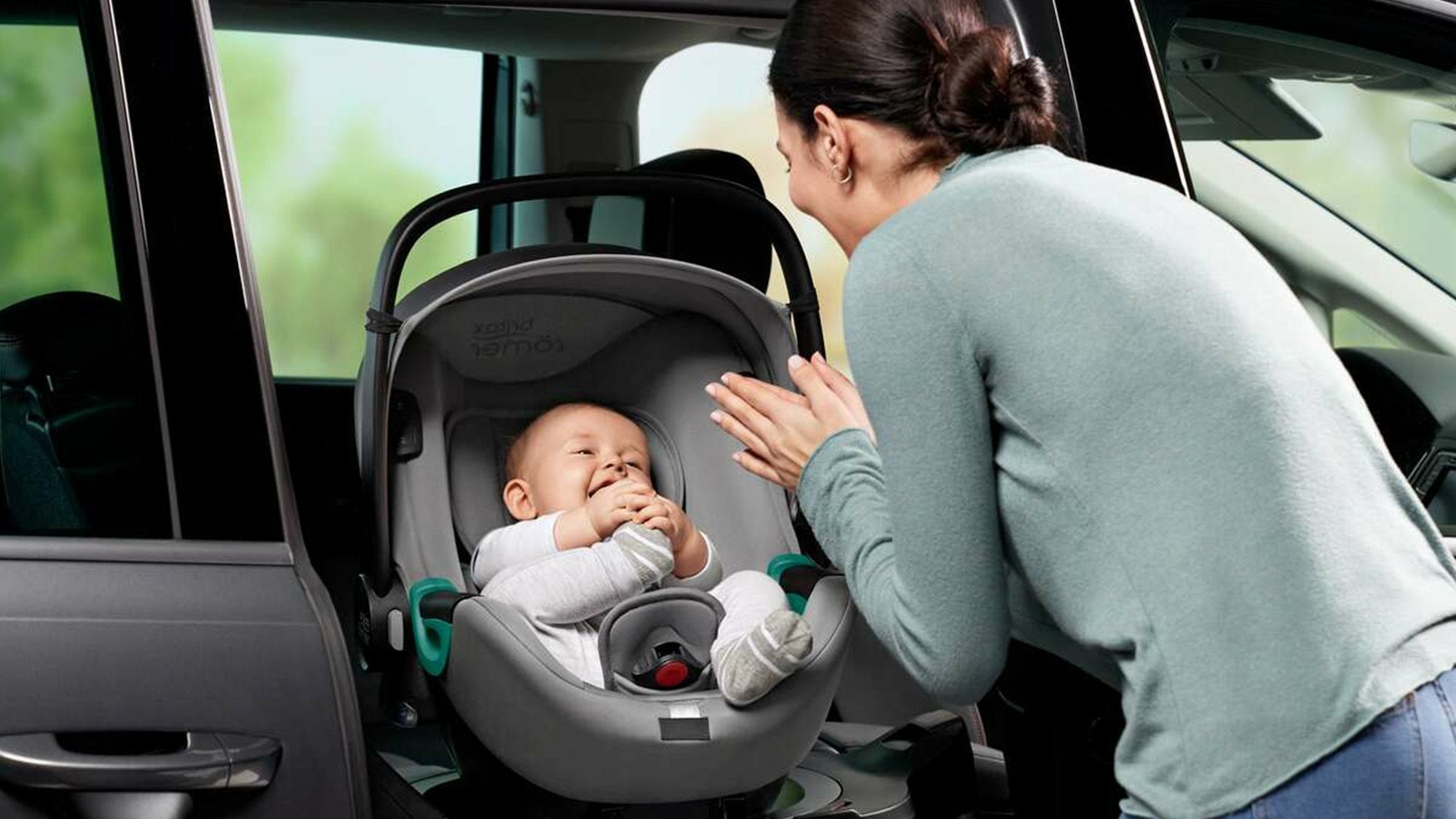 Happy baby in child seat in car Mother stands in front and claps