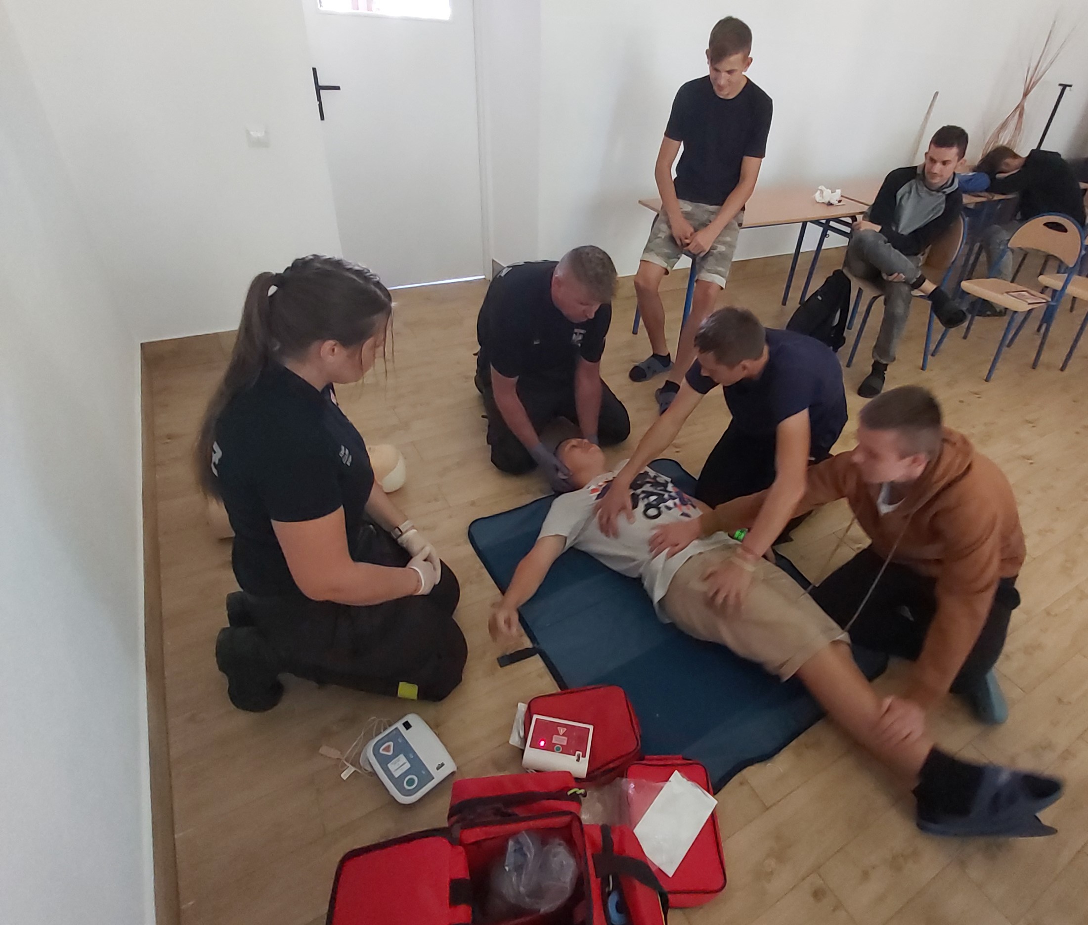 Warsztaty z pomocy przedmedycznej | Wychowankowie MOW ćwiczą na sobie ukłądanie poszkodowanego w pozycji bocznej, obok Druhowie tłumaczą jak przeprowadzić ćwiczenie, z tyłu przyglądają się kolejni uczniowie..jpg