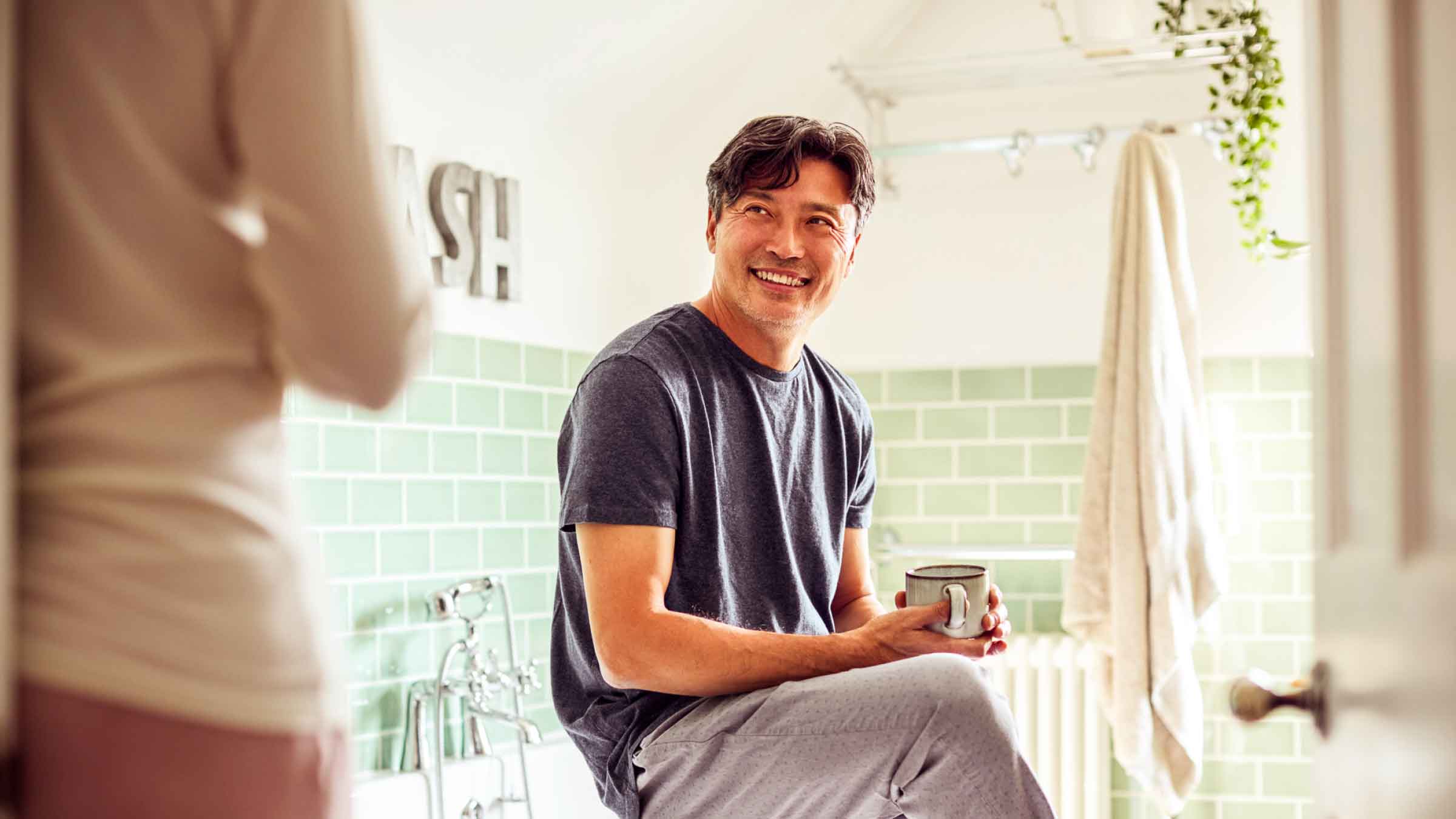 Man perched on side of bathtub holding his morning coffee cup while smiling at his partner who's standing in the doorway.